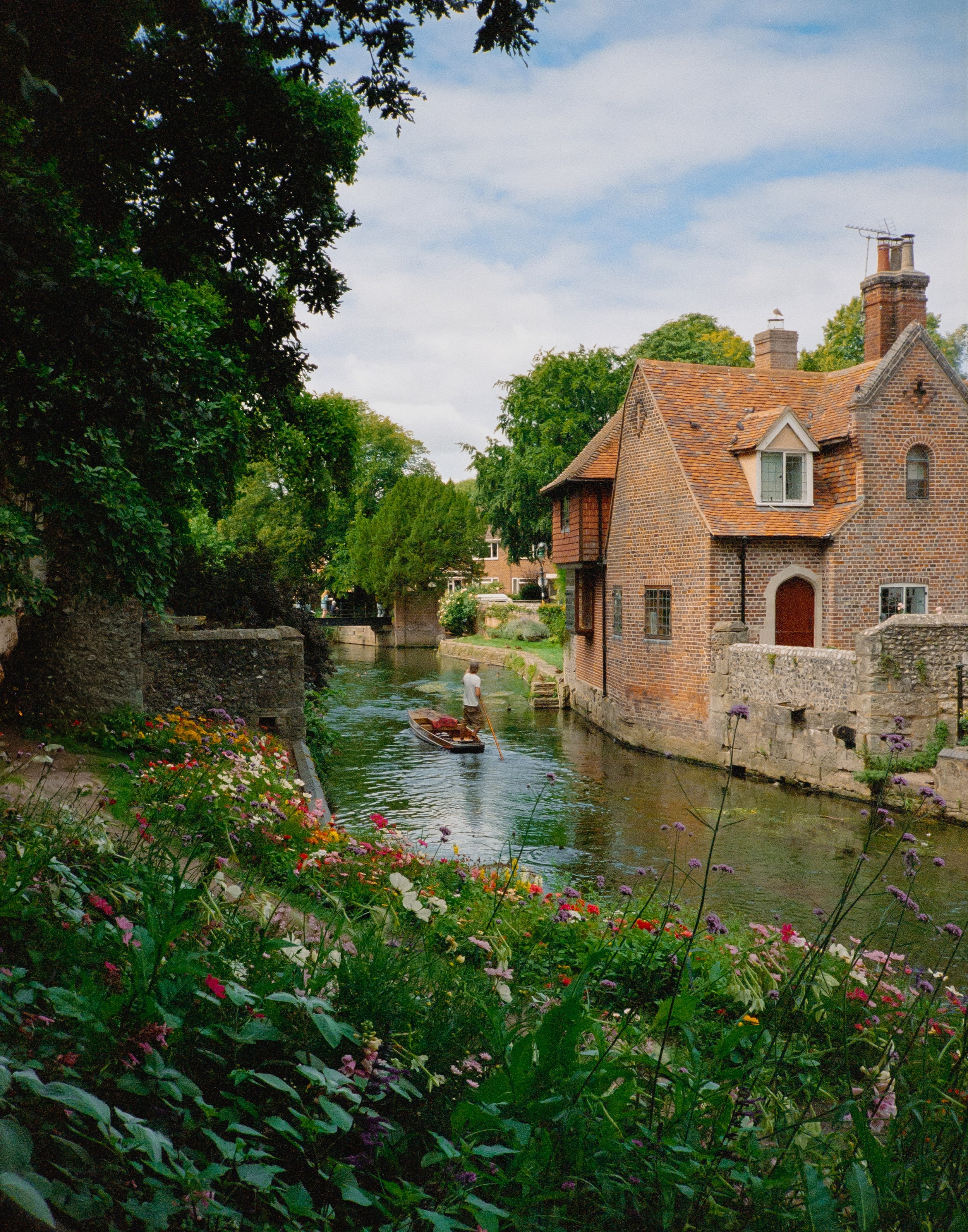 A person rowing a boat in a small canal with a brick house on the water's edge, surrounded by greenery and colorful flowers.
