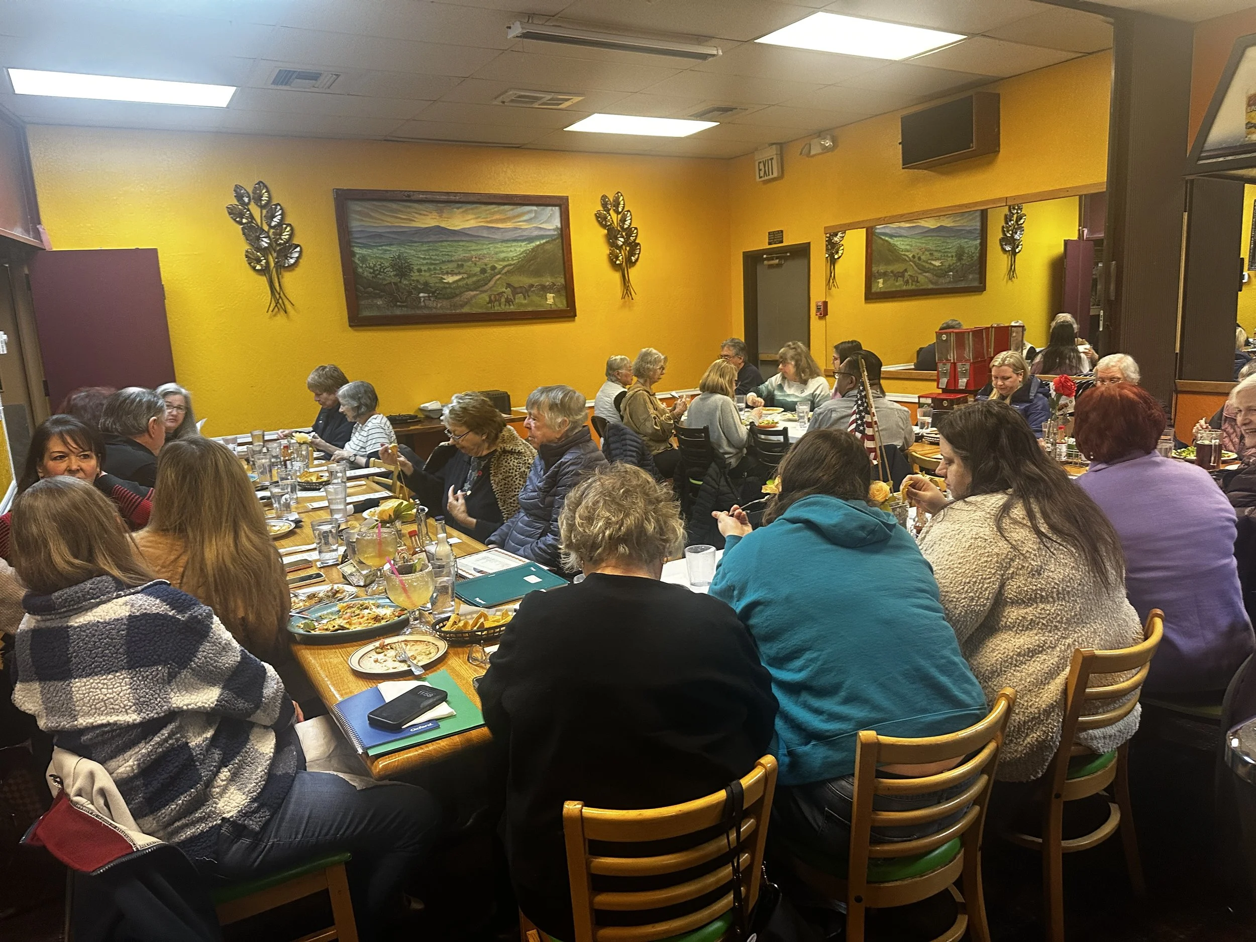 A large group of people gathered around dining tables in a restaurant with yellow walls, mountain landscape paintings, and decorative wall art, enjoying a meal together.