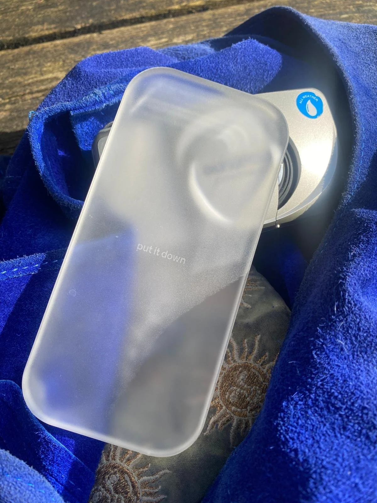 A frosted glass phone snuggled in a blue fleece blanket, with a camera and a sticker labeled 'All Weather.'