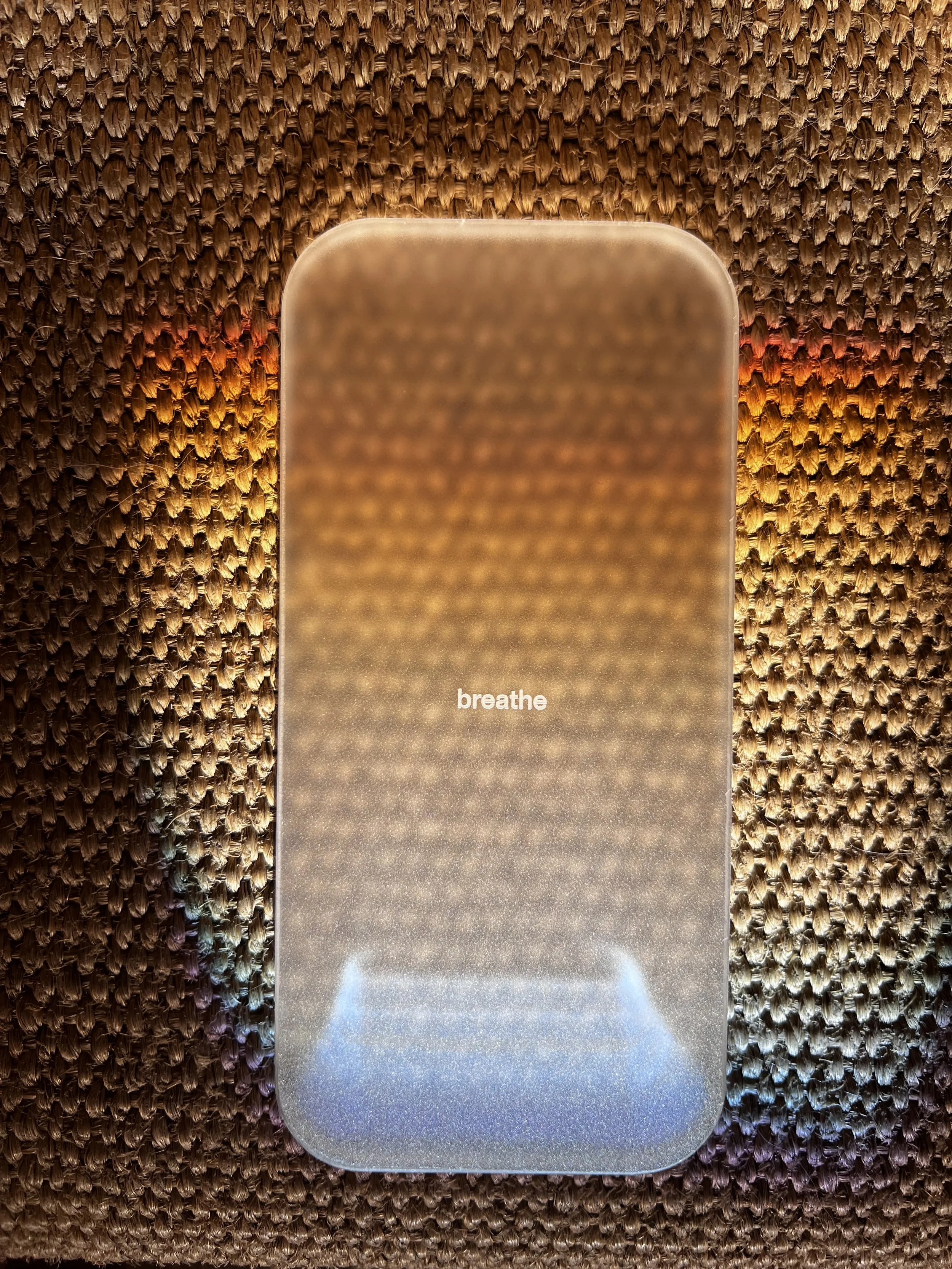 A smartphone with a reflective screen displays the word 'breathe' in the center. The phone is placed on a textured, woven surface with warm lighting.