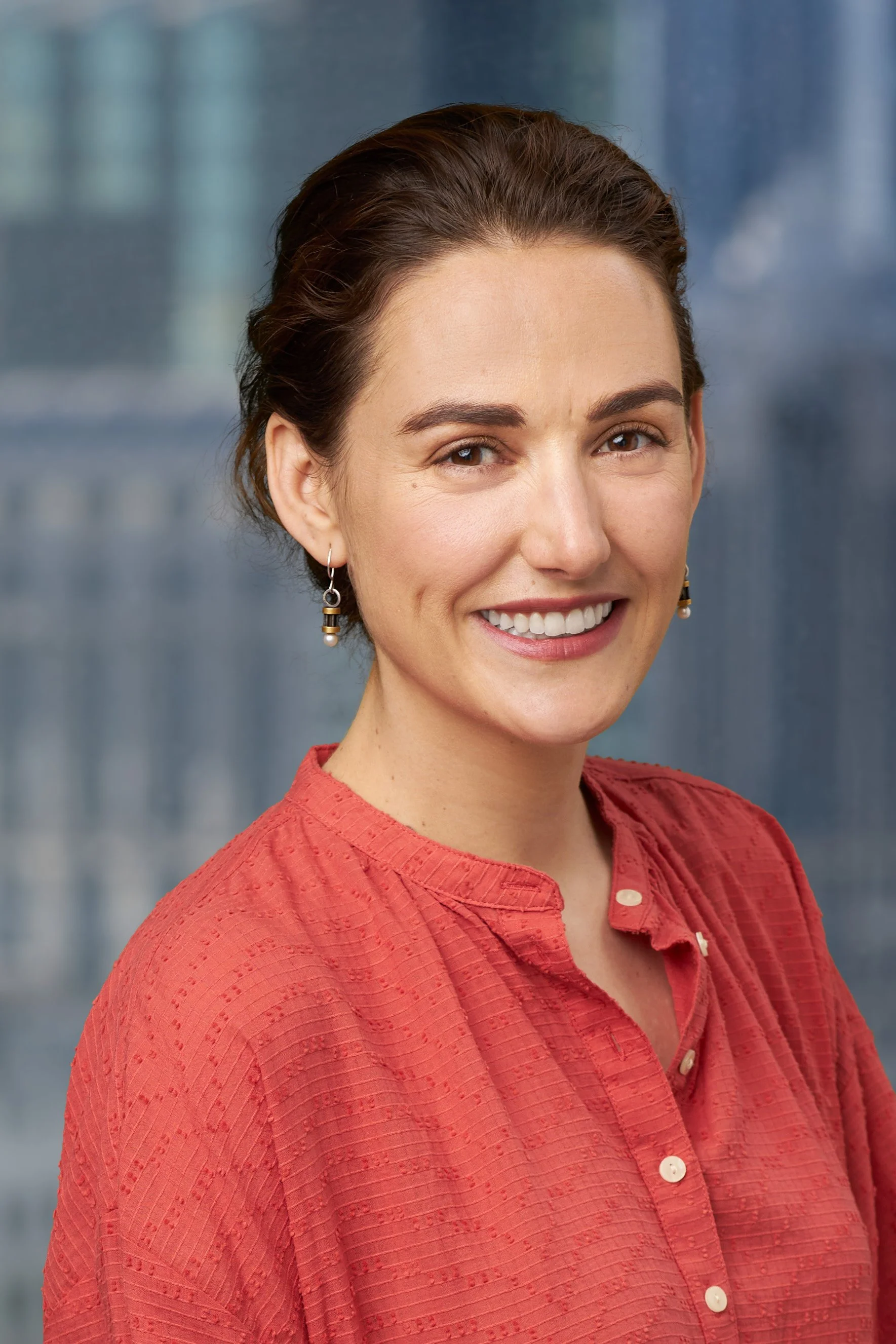A woman with short brown hair, brown eyes, and a bright smile, wearing earrings and a red textured button-up shirt, standing against a blurred blue background.