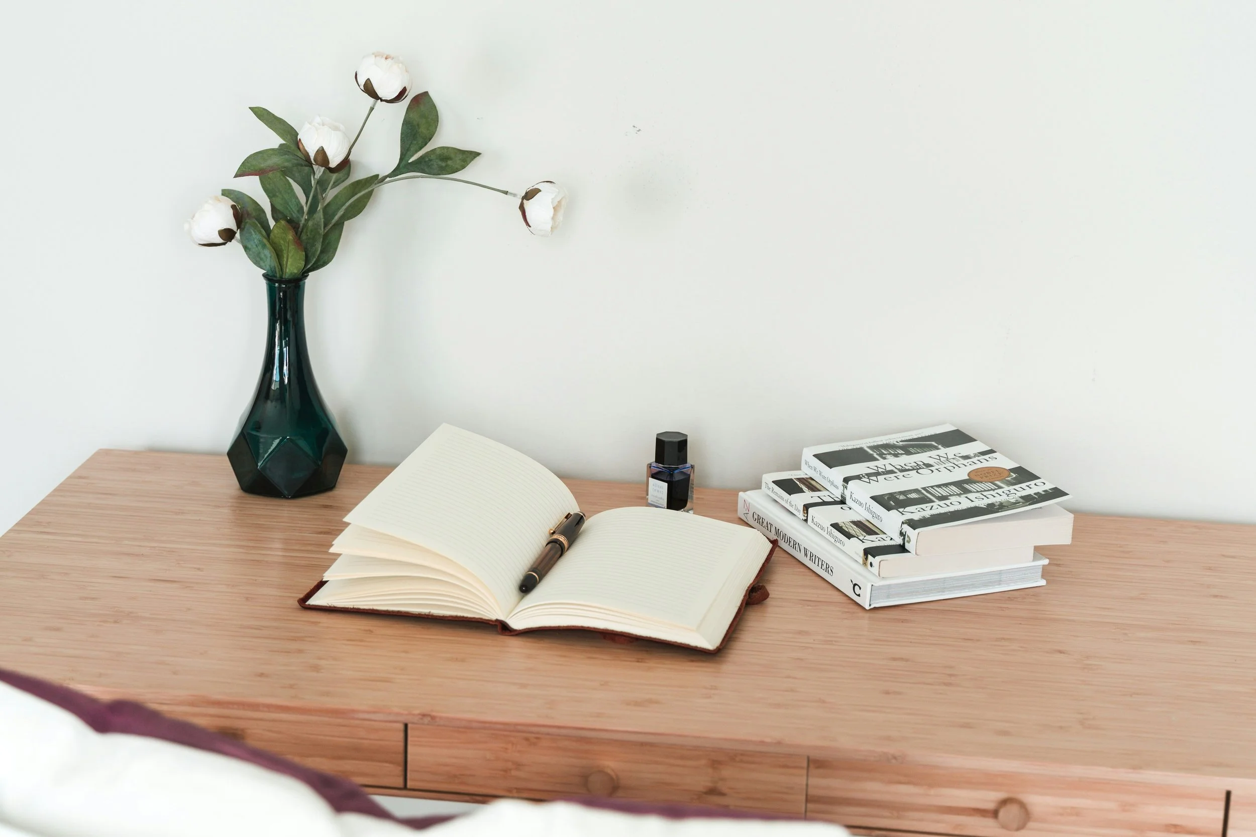 A wooden desk with an open notebook, a black pen, a small bottle of ink, a stack of books, and a dark green vase with cotton flowers and green leaves against a white wall.