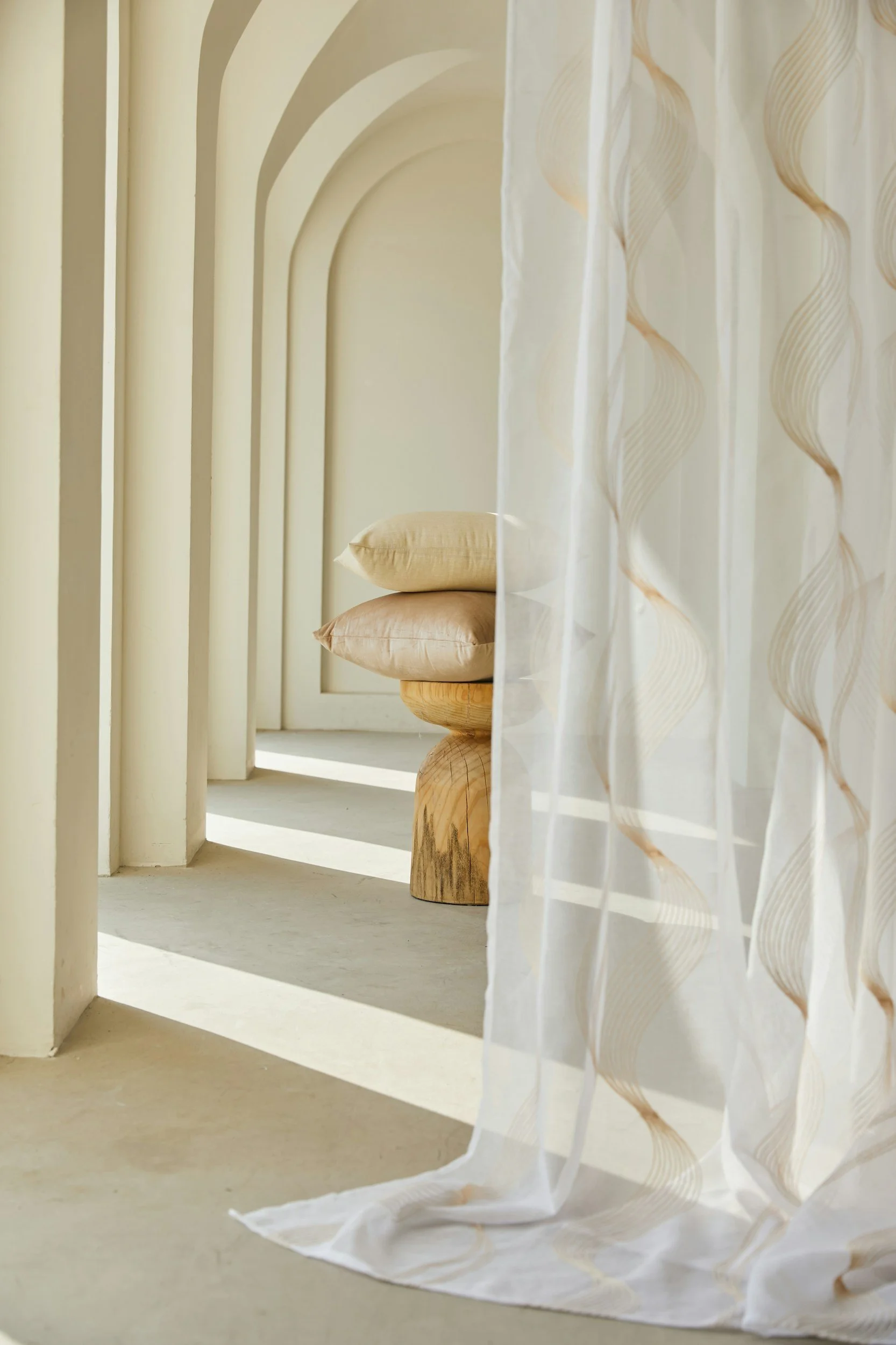 Soft beige pillows stacked on a wooden stool next to a sheer curtain with a wavy pattern, sunlight casting shadows through a series of archways.