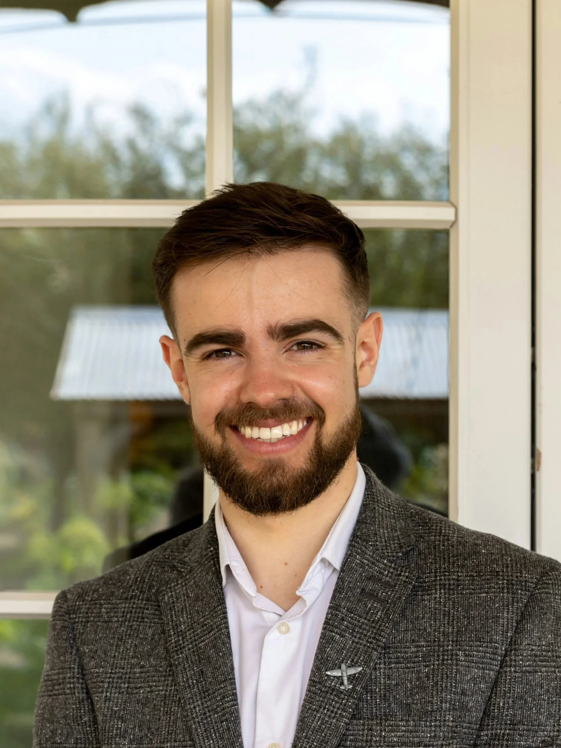 A smiling man in a gray blazer and white shirt standing in front of a window with a view of trees outside.