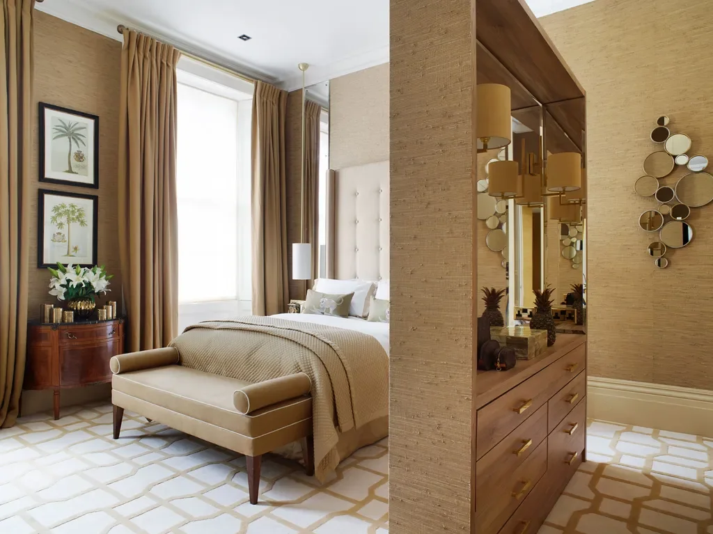 A luxurious hotel bedroom divided by a wooden and mirror partition wall. The room features a bed with white linens, beige bedding, and a beige upholstered bench at the foot. There are large beige curtains, framed botanical prints, a wooden side table with white flowers, and a dresser with decorative pineapple jars. The walls have a textured beige finish, and the room is bright with natural light.