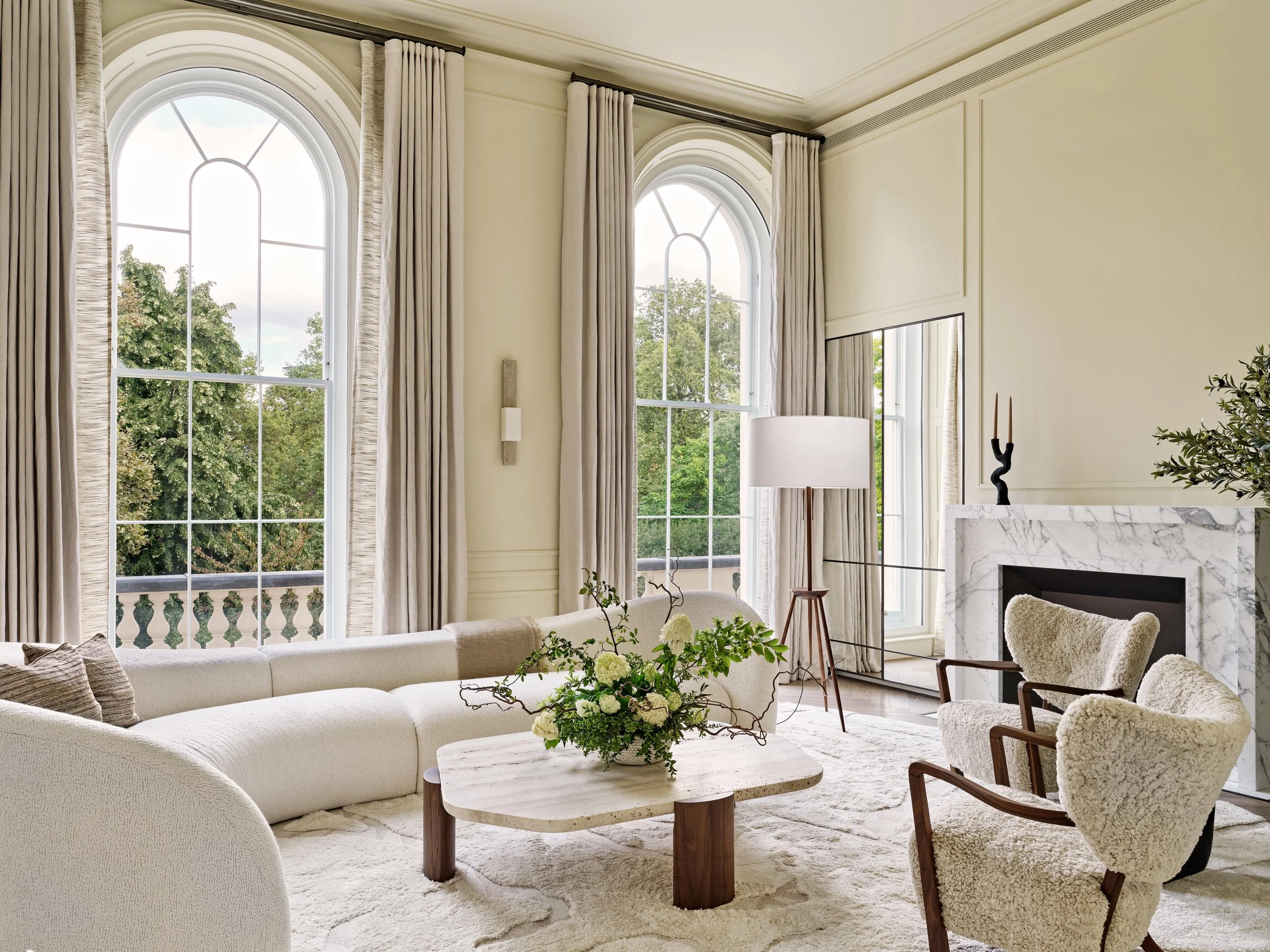 Bright living room with large arched windows, cream-colored curtains, contemporary white sofa, two armchairs with wooden frames and plush upholstery, a marble fireplace, a tall floor lamp, and a floral arrangement on a marble coffee table.