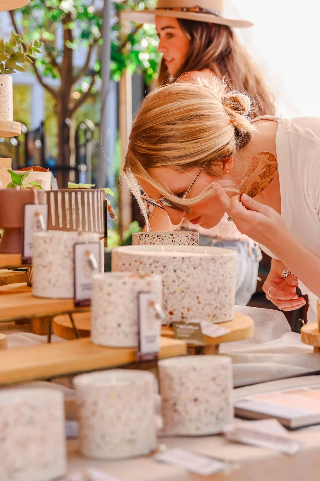 A woman with blonde hair, glasses, and a tattoo on her shoulder examines a speckled cylindrical container at an outdoor market, with another woman wearing a hat in the background.