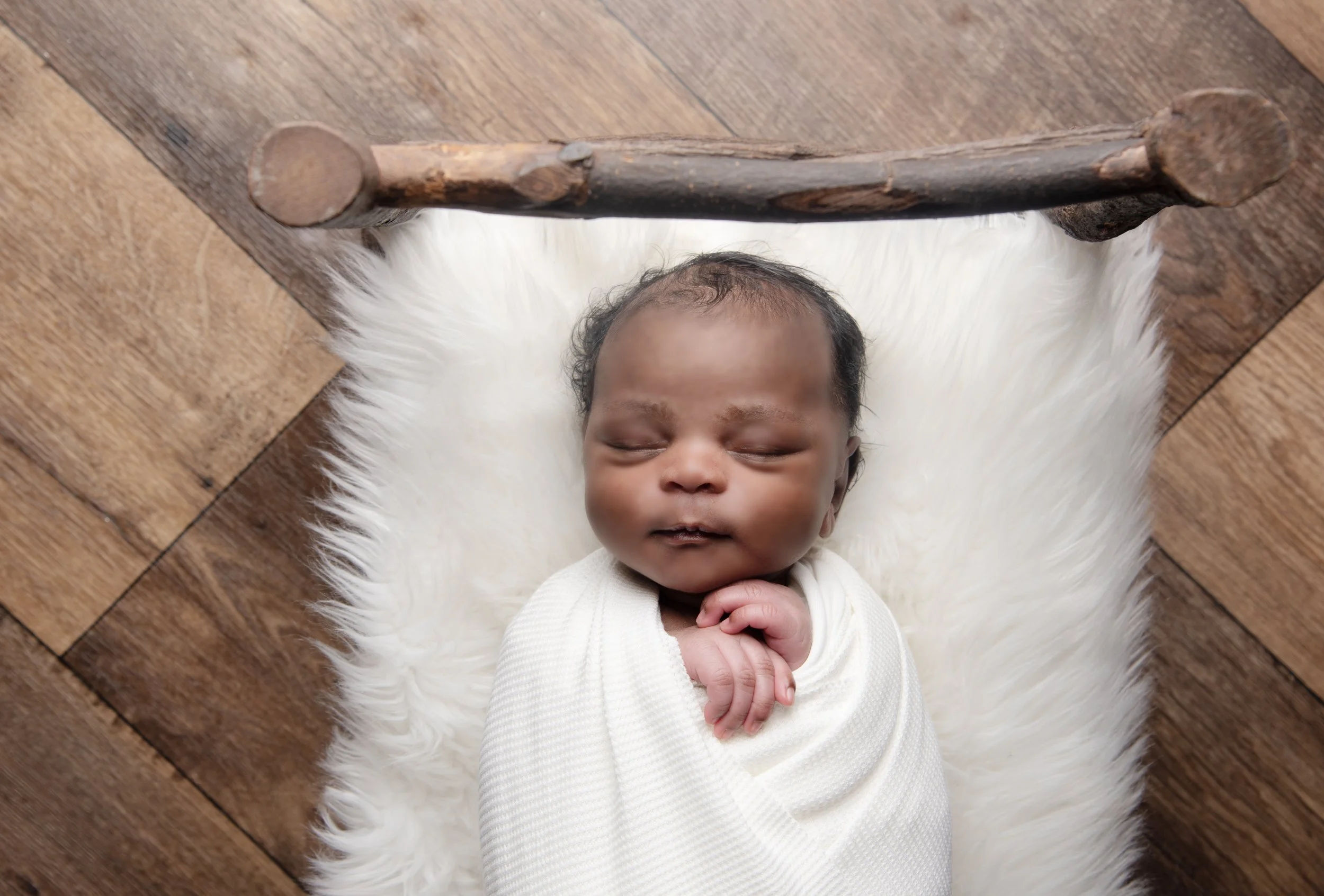 A sleeping baby swaddled in white fabric, lying on a fluffy white rug, with a wooden stick propped above the baby's head, on a wooden floor.