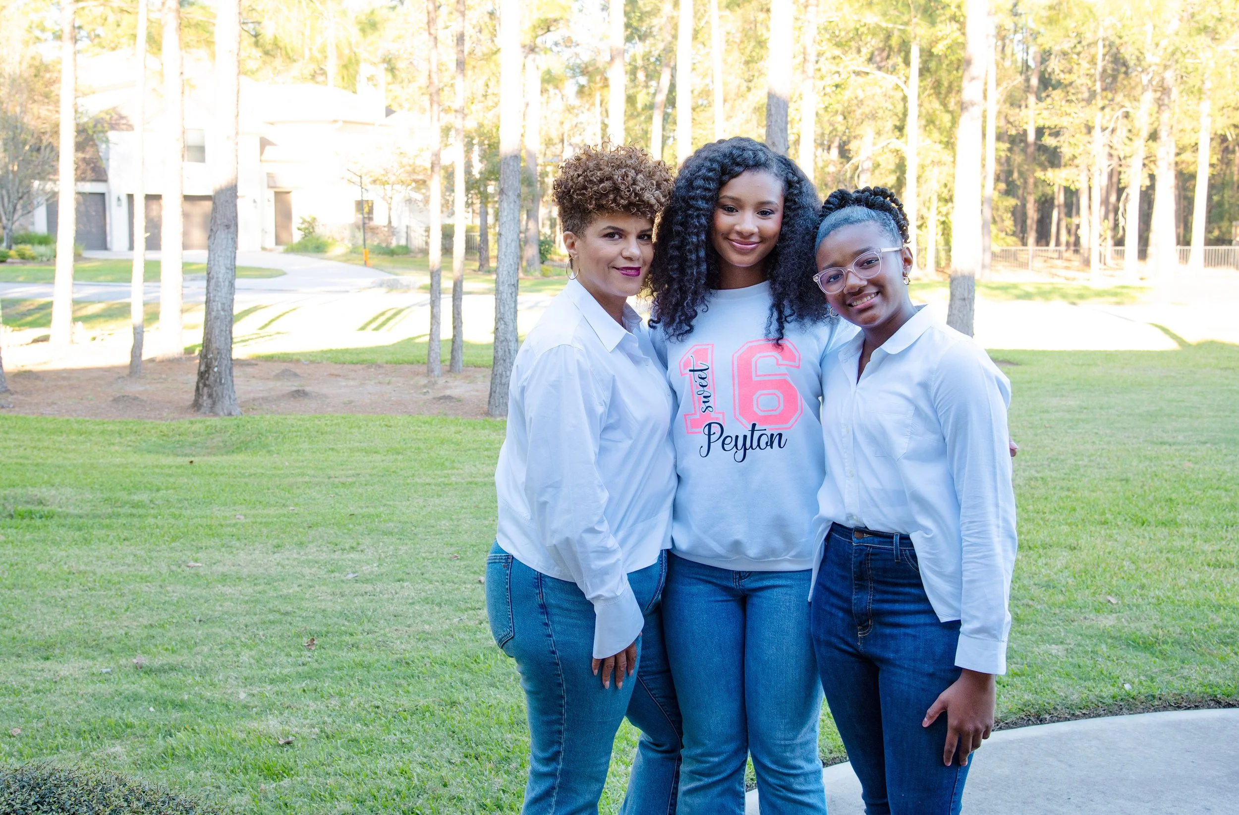 Three women standing together outdoors on a grassy area with trees and houses in the background, smiling and posing for a photo.