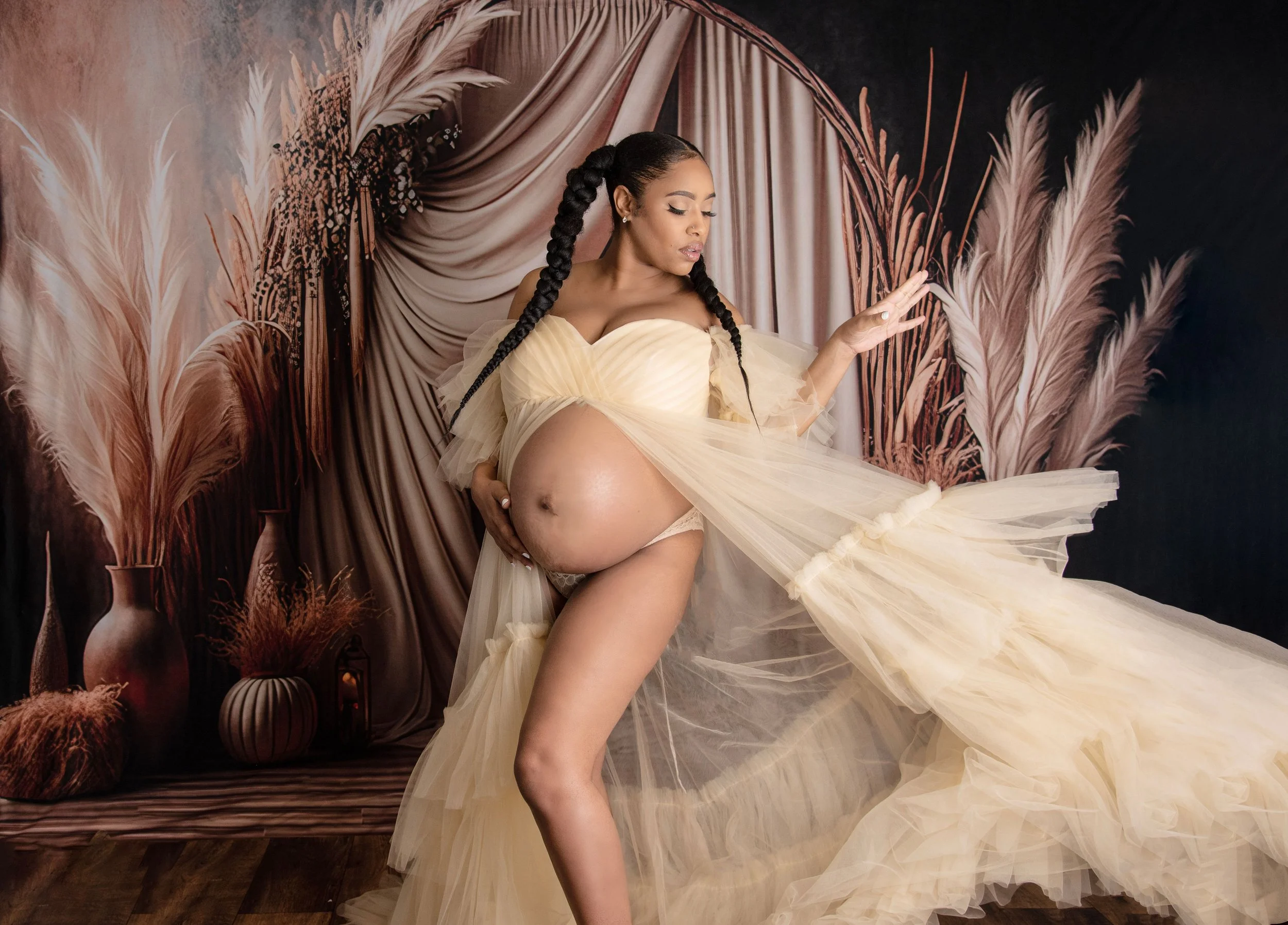 A pregnant woman with long braids wearing a flowing cream-colored gown, standing in front of a backdrop of pink and beige pampas grass and draped fabric.