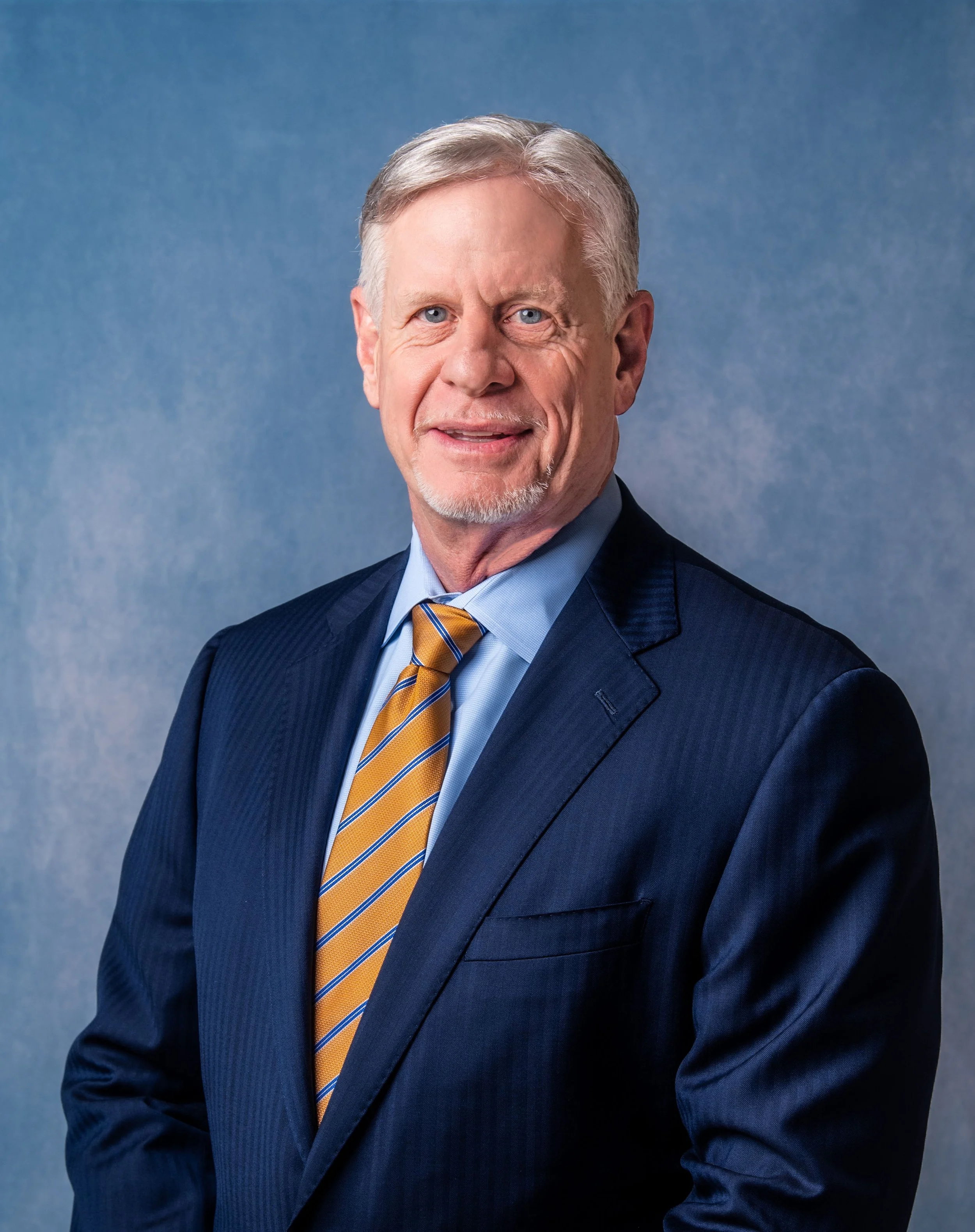 A professional headshot of a man with gray hair, blue eyes, and a goatee, wearing a navy blue suit, light blue shirt, and striped yellow and blue tie, standing against a blue textured background.