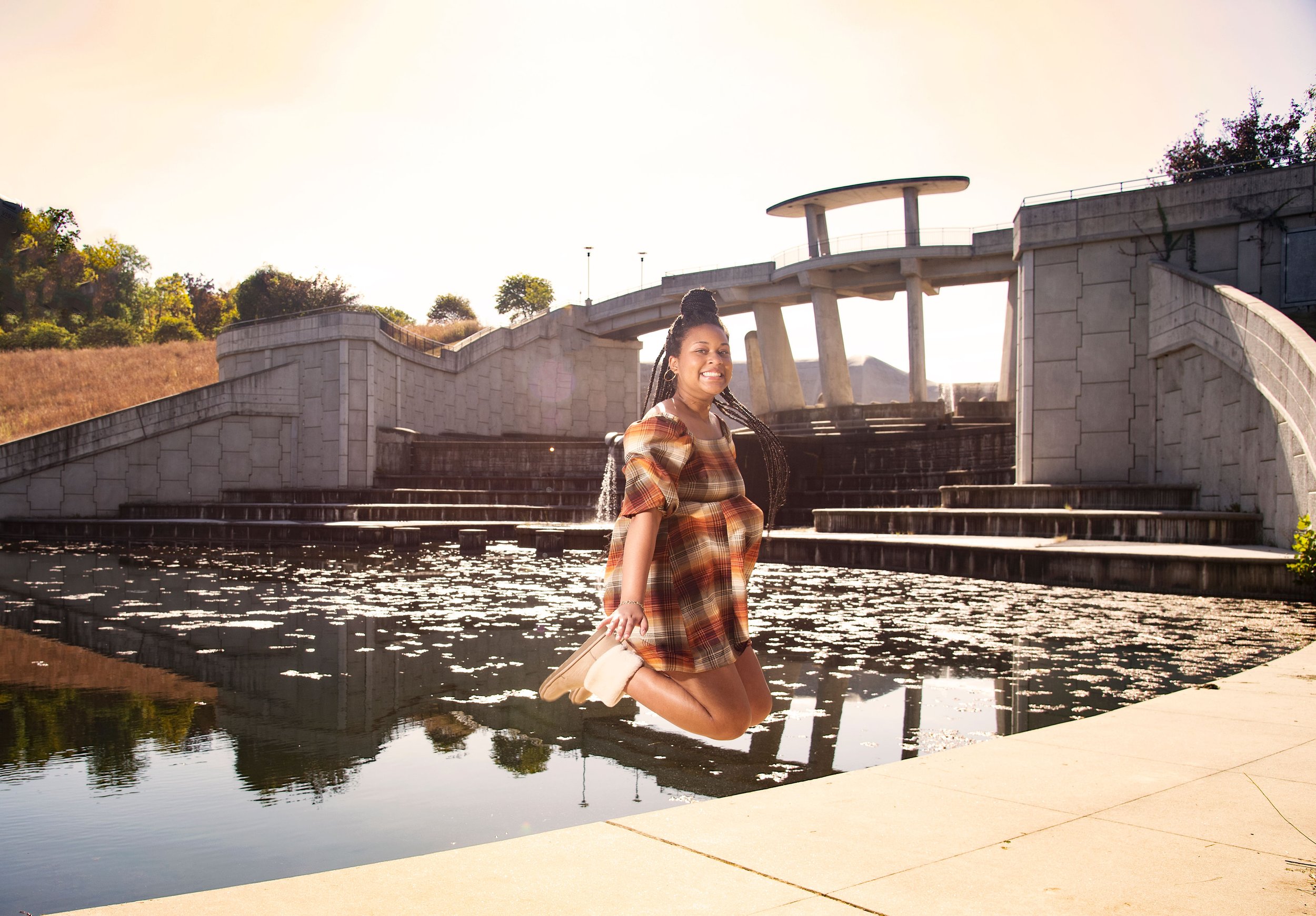 A woman in a plaid dress is jumping over a reflecting pool in an outdoor park or urban space with modern concrete structures and greenery in the background.