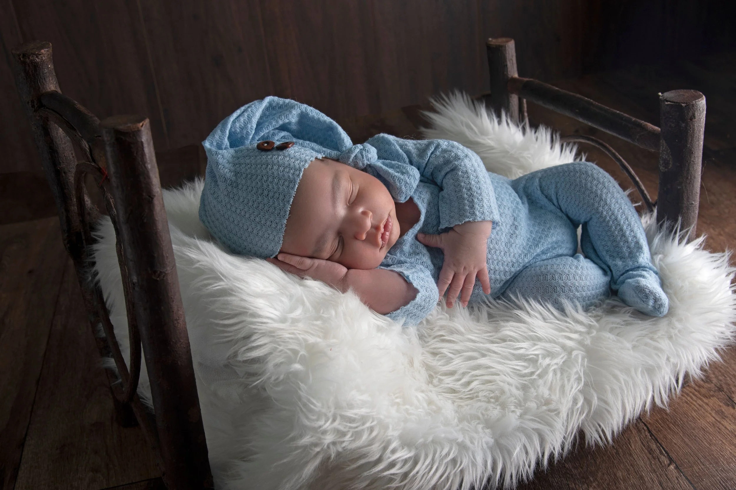 A sleeping newborn baby in a blue knitted outfit and matching hat, lying on a fluffy white fur blanket on a small rustic bed.