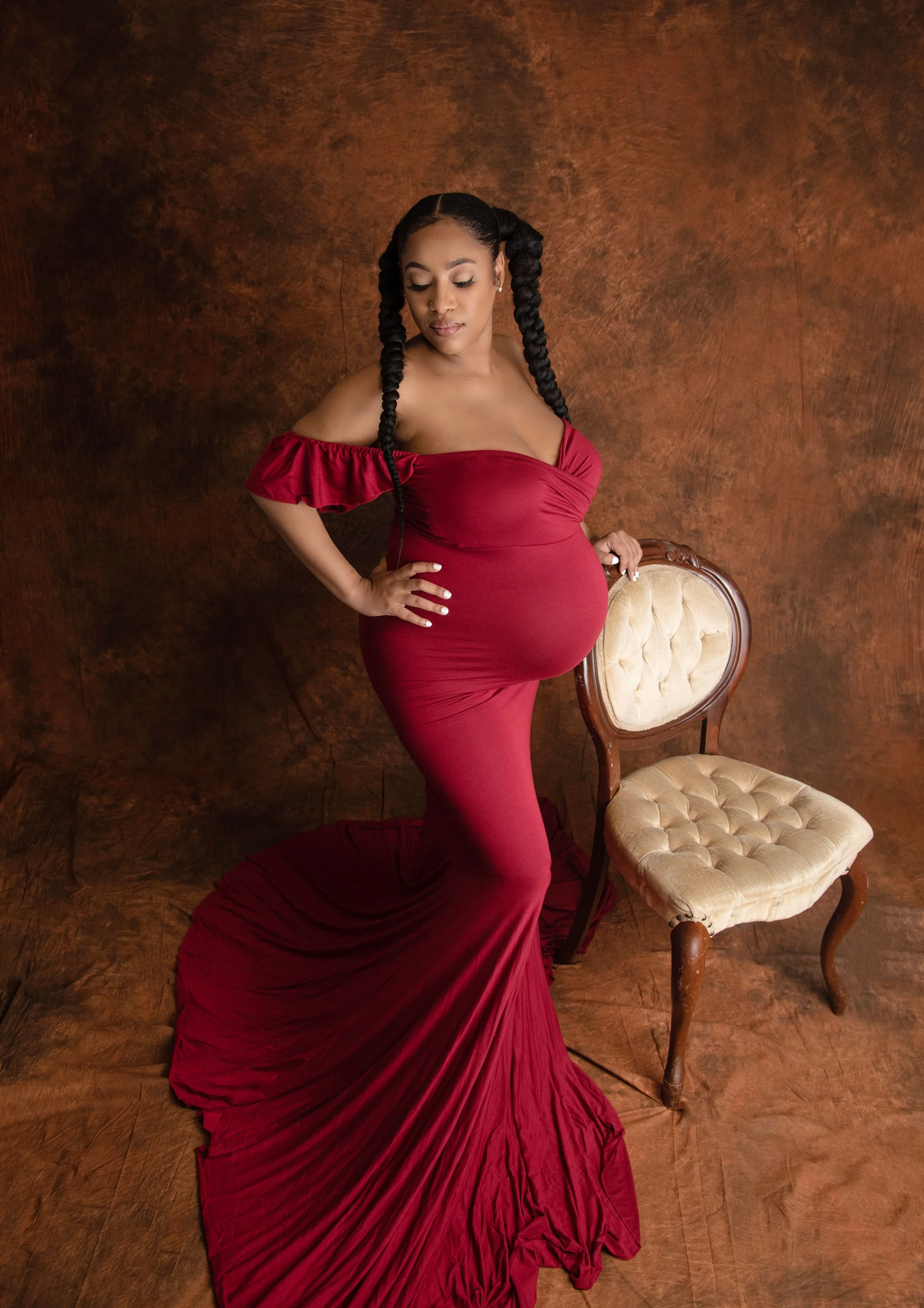 A pregnant woman in a fitted, off-the-shoulder red gown poses next to a vintage beige and wood chair against a textured brown backdrop.