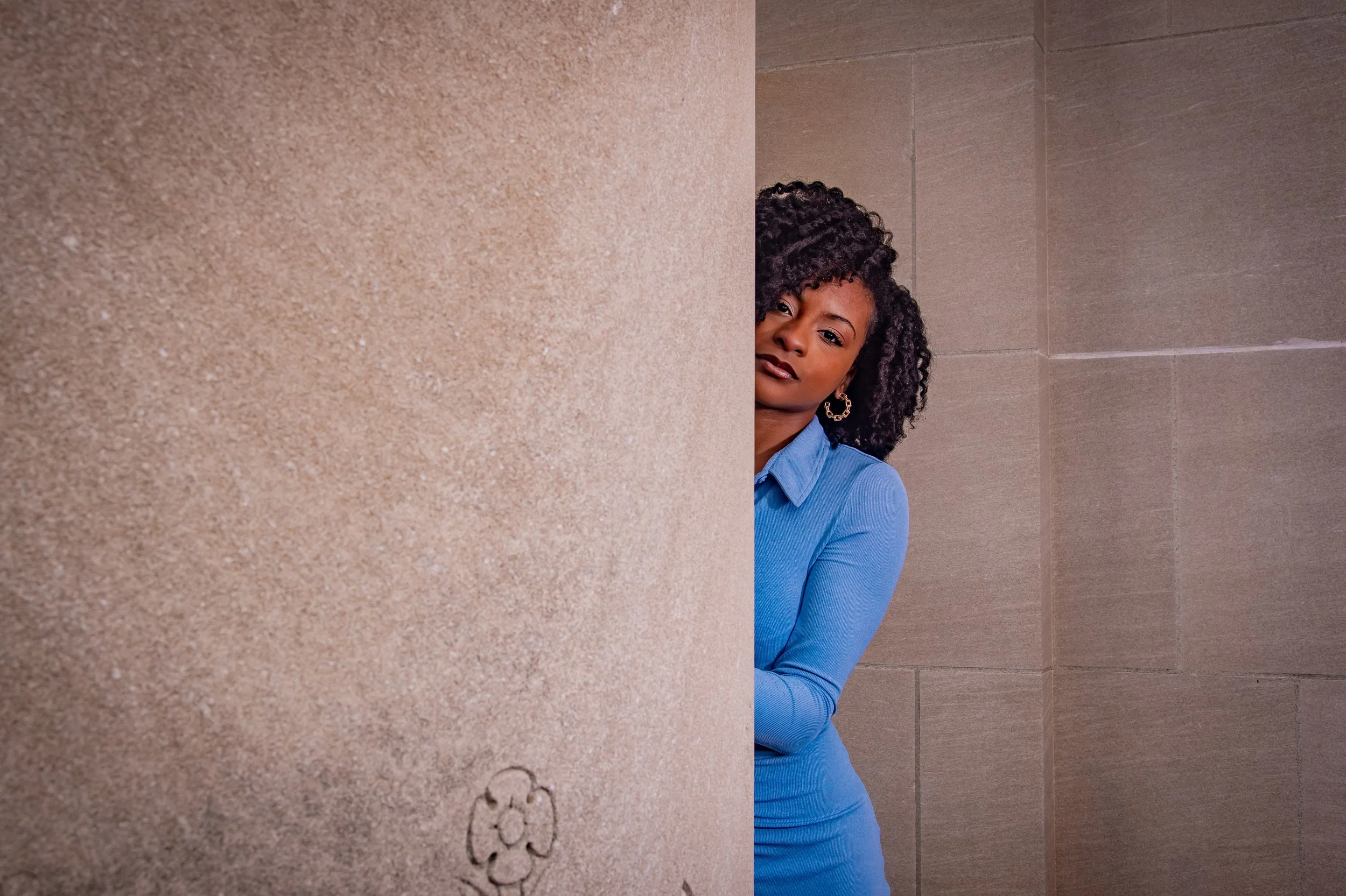 A woman with curly black hair wearing a blue shirt, peeking out from behind a beige wall.