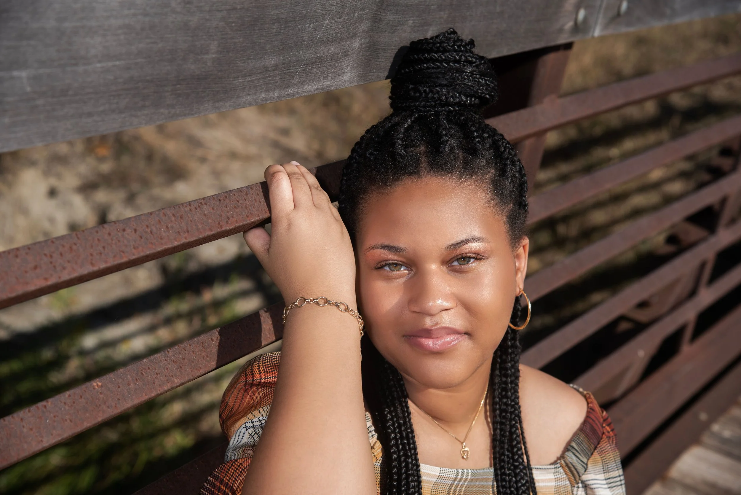A young woman with dark braided hair styled in a high bun, wearing hoop earrings and a necklace, sitting on a wooden and metal bench outdoors. She is looking at the camera with a slight smile, resting her hand on her head.