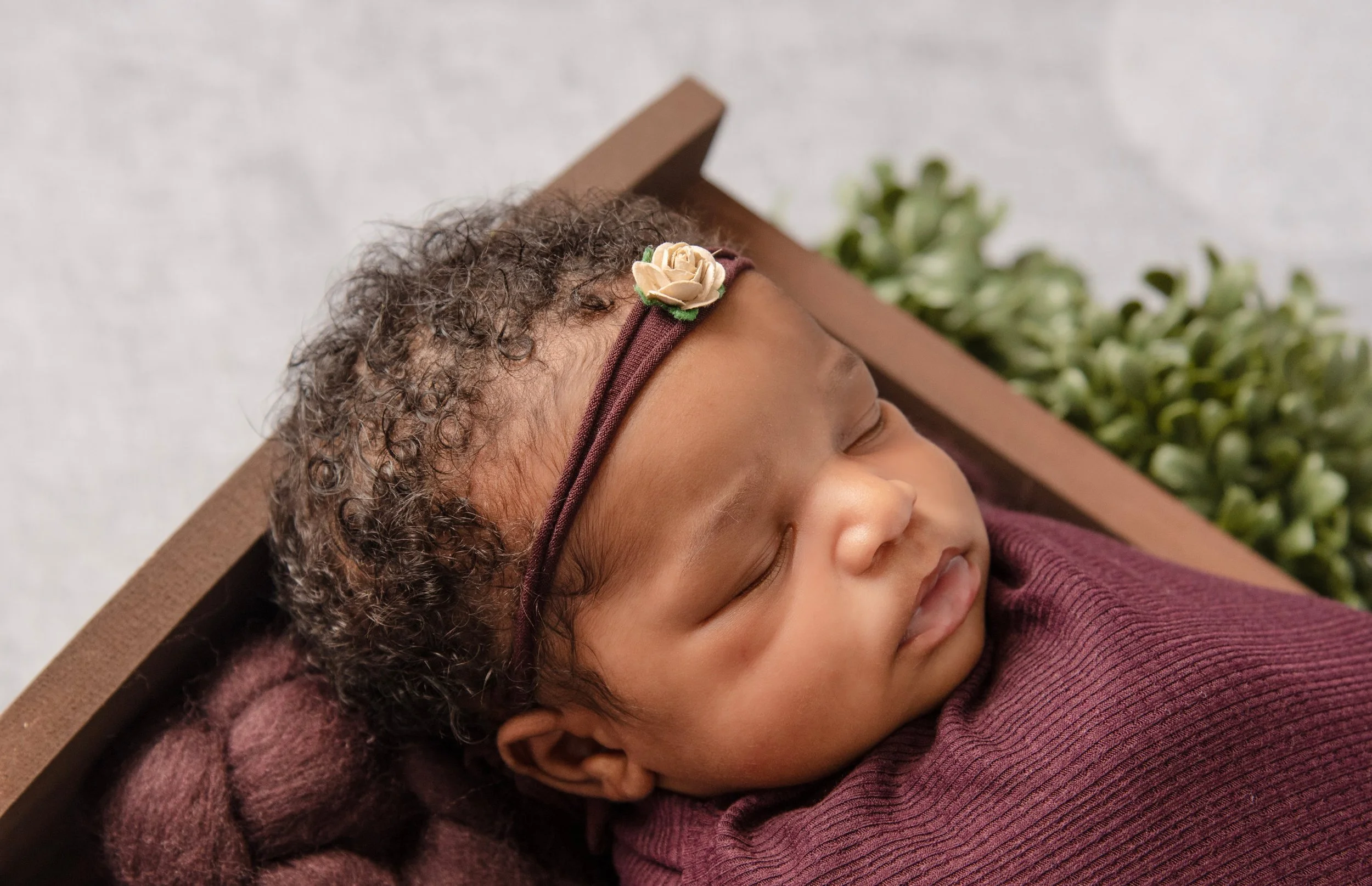 Close-up of a sleeping baby with curly hair, wearing a brown headband with a small flower, wrapped in a maroon blanket, and resting on a brown pillow with greenery in the background.