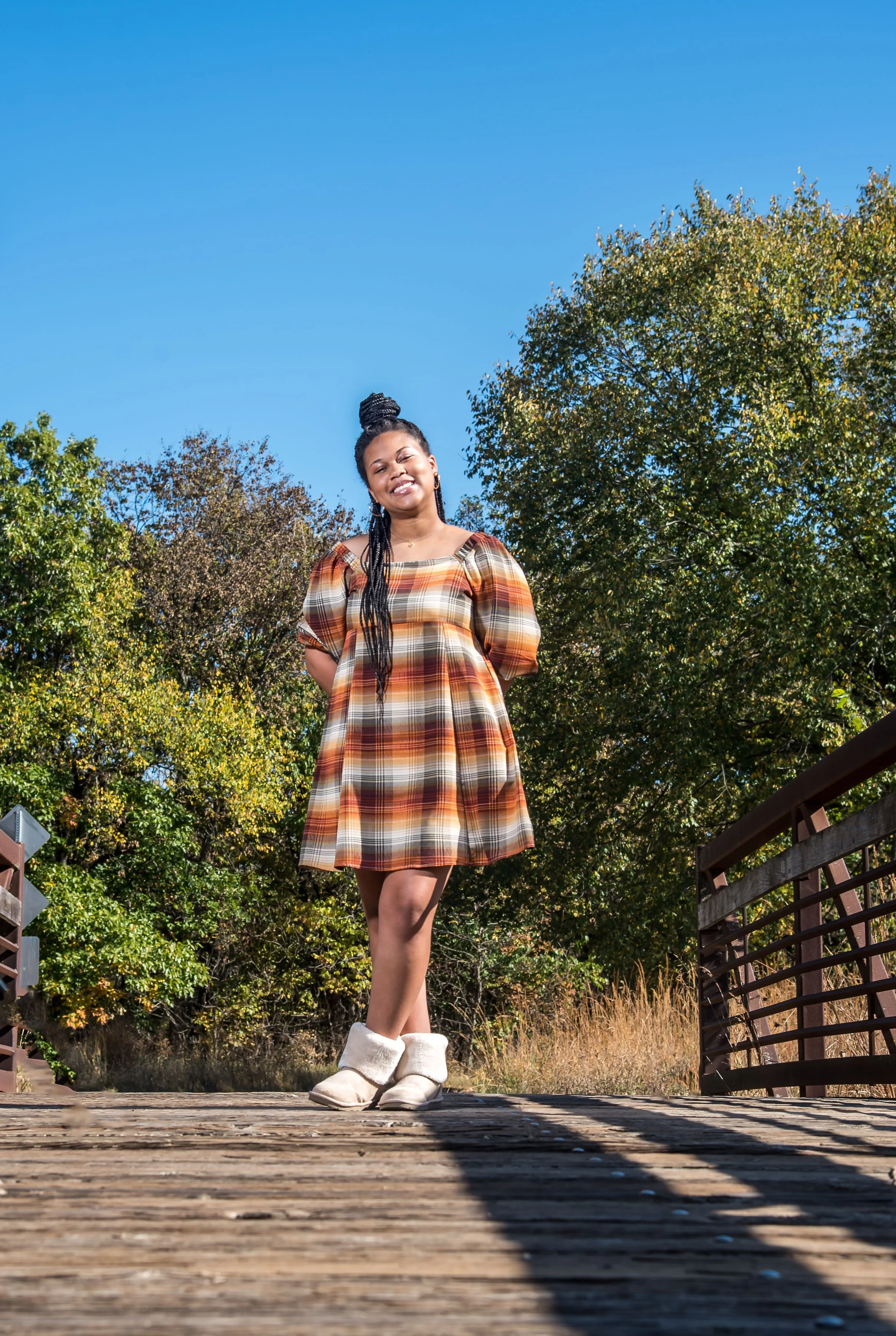 Young woman smiling and standing on a wooden bridge outdoors with green trees and a clear blue sky in the background.