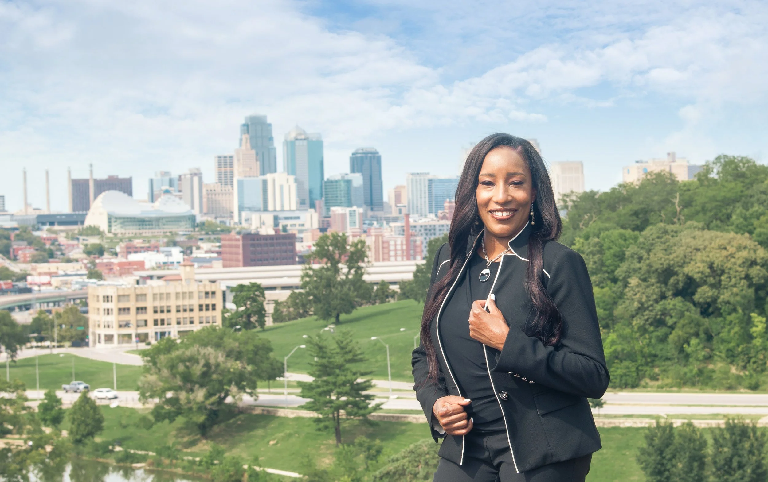 A smiling realtor in a black suit with white piping, standing outdoors with a city skyline and green park behind her.