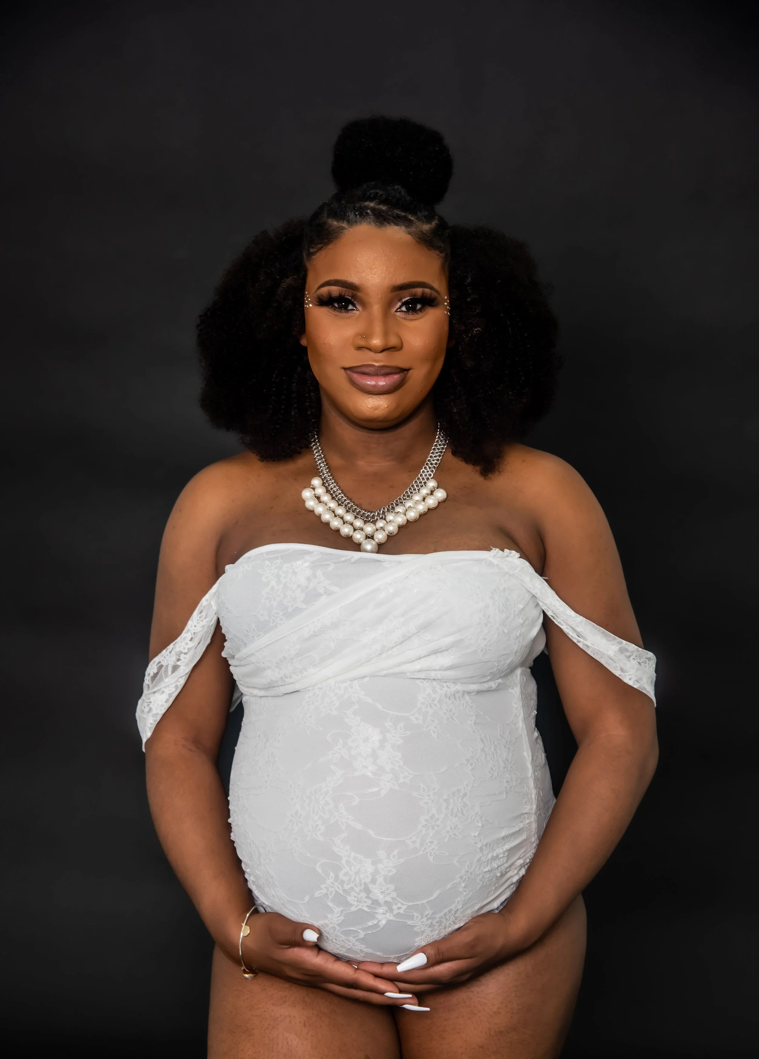 Expecting mom smiling into the camera wearing a white lace bodysuit while cradling her baby bump