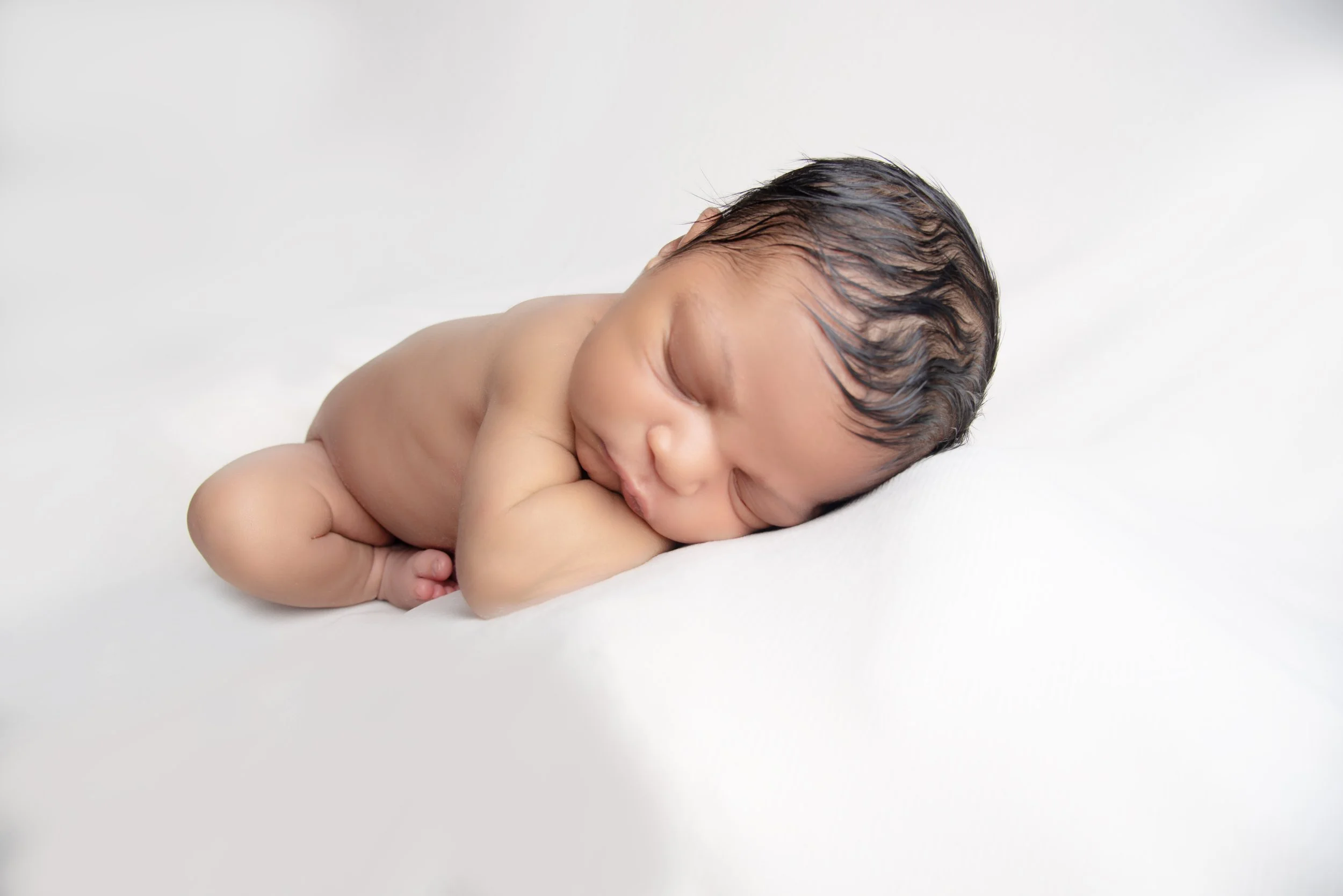 A newborn baby sleeping on a white surface, curled up on their side with head resting on arms and legs tucked in.