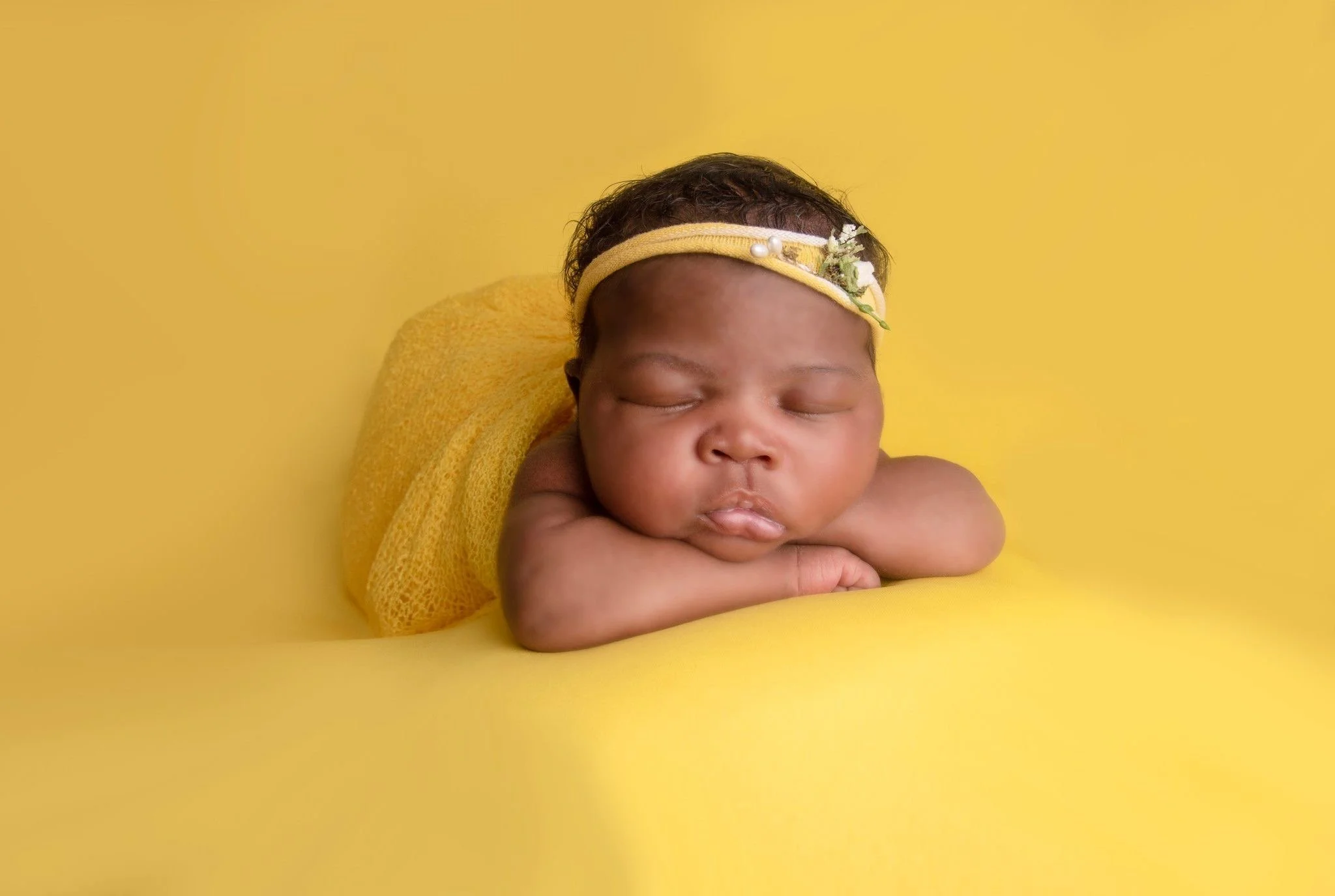 Newborn baby girl wrapped in yellow fabric against yellow fabric wearing a yellow headband