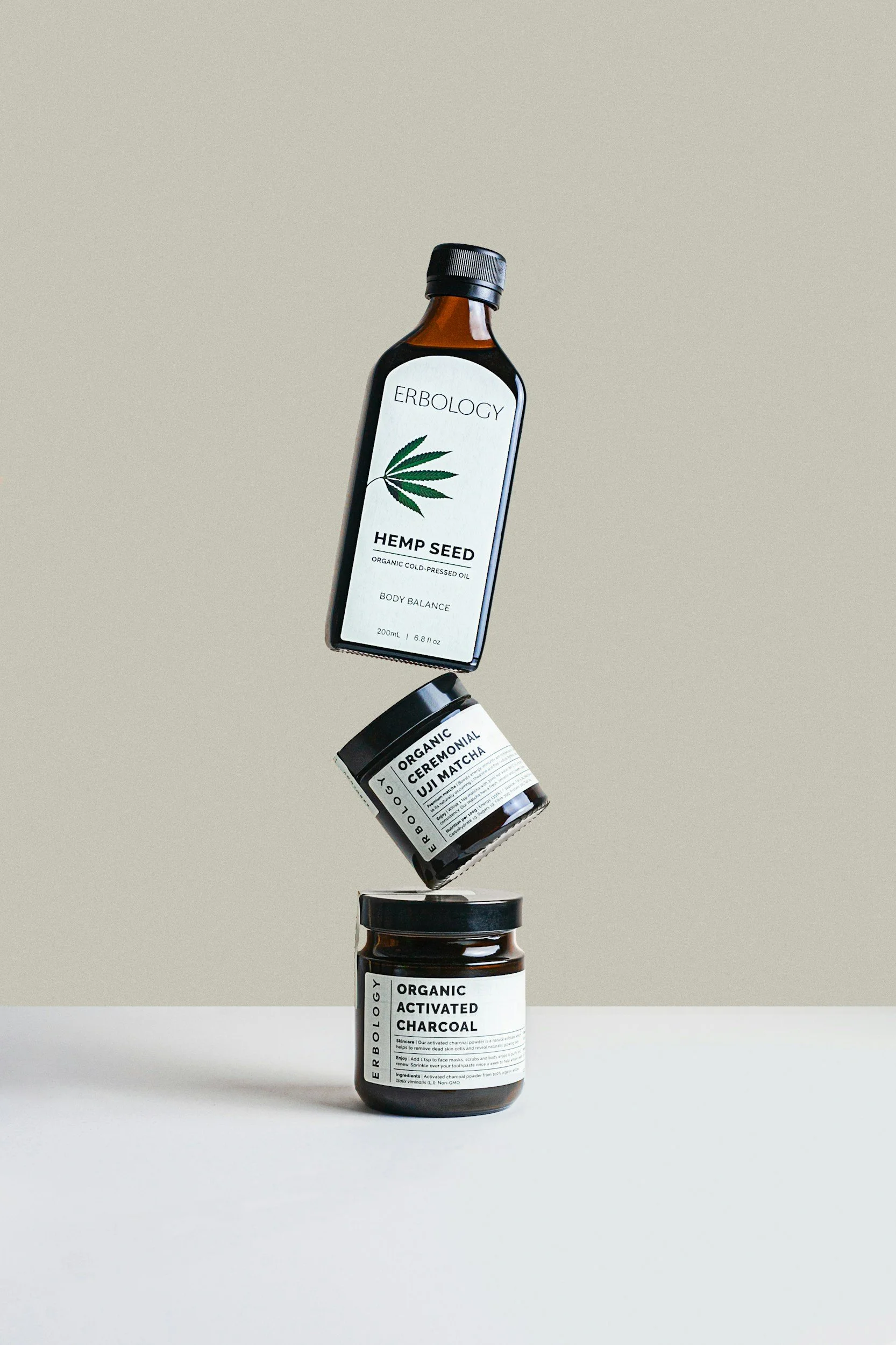 Three health product containers in mid-air with a minimalist background: a bottle of Erbology hemp seed oil, a jar of organic ceremonial Uji matcha, and a jar of organic activated charcoal.