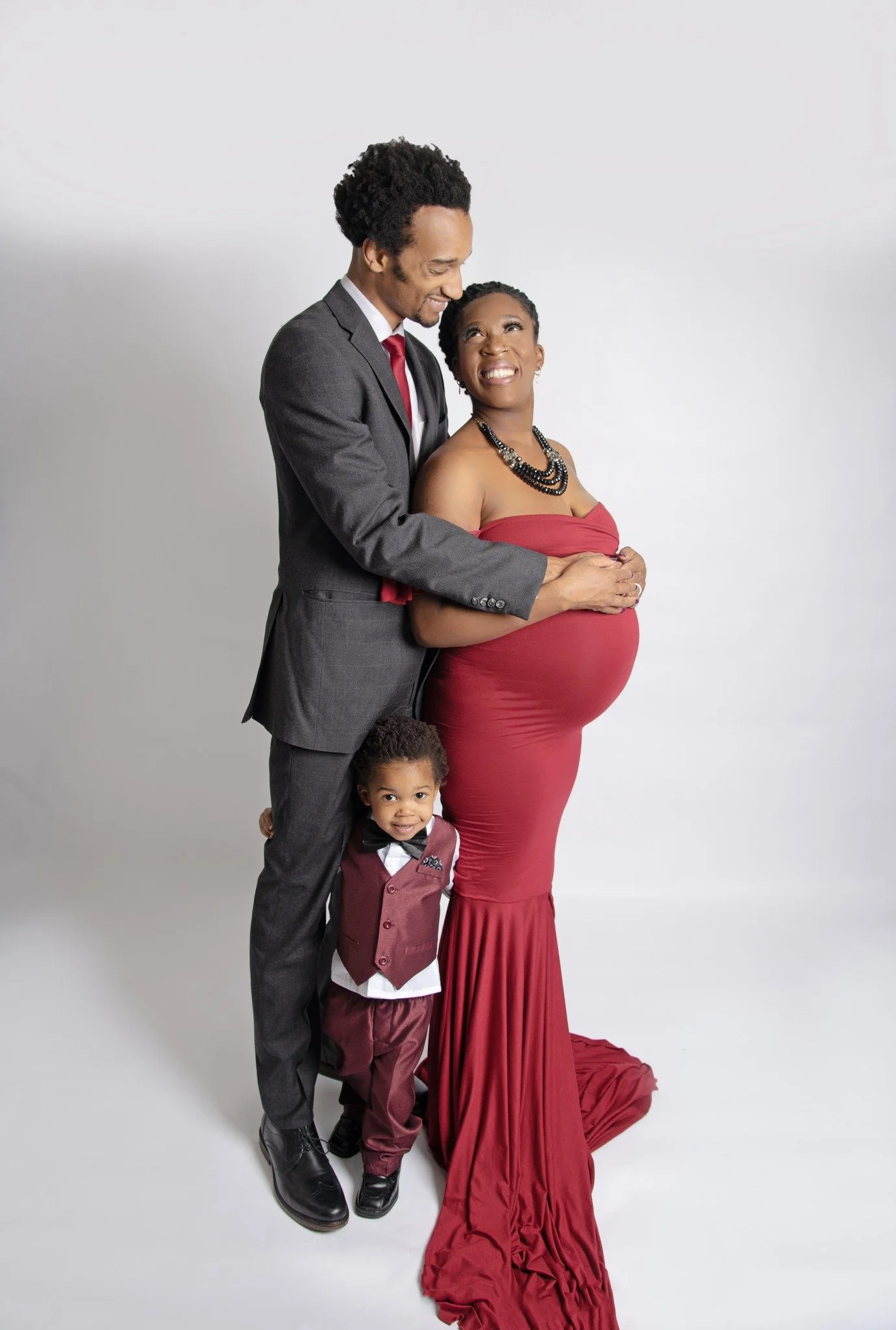 A family of three, a pregnant woman in a red dress, a man in a suit, and a young child in matching maroon and white attire, posing together against a white background.
