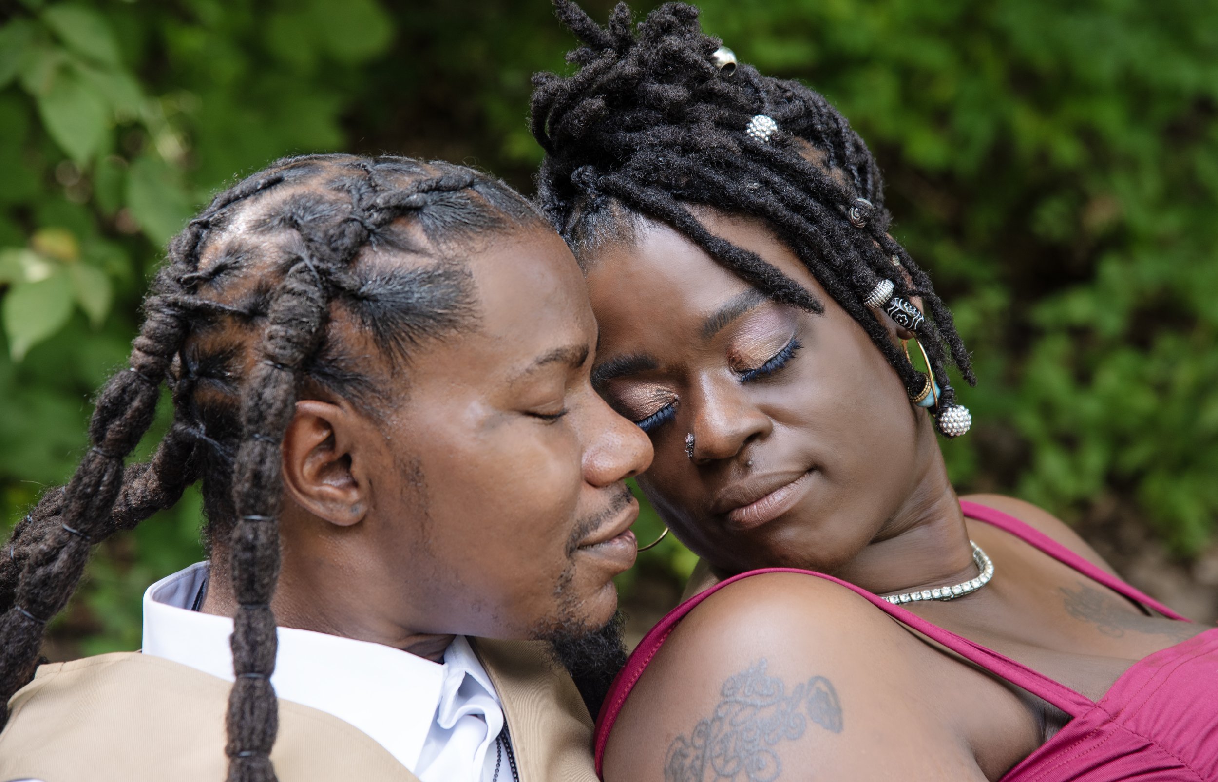A close-up of a couple with their foreheads touching, eyes closed, outdoors with green foliage in the background.