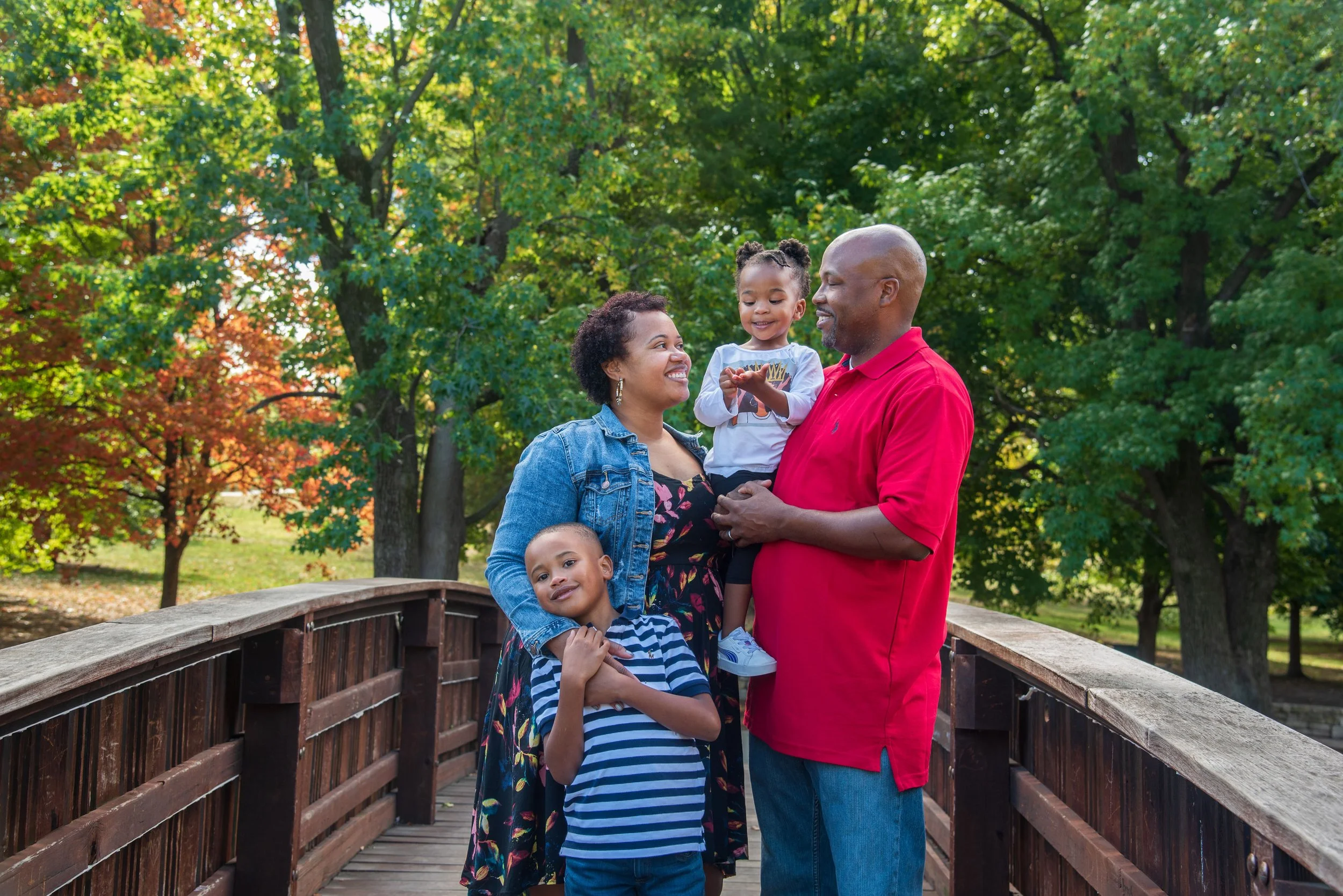 A family of four standing on a wooden bridge in a park with green trees and colorful foliage, smiling and enjoying quality time together.