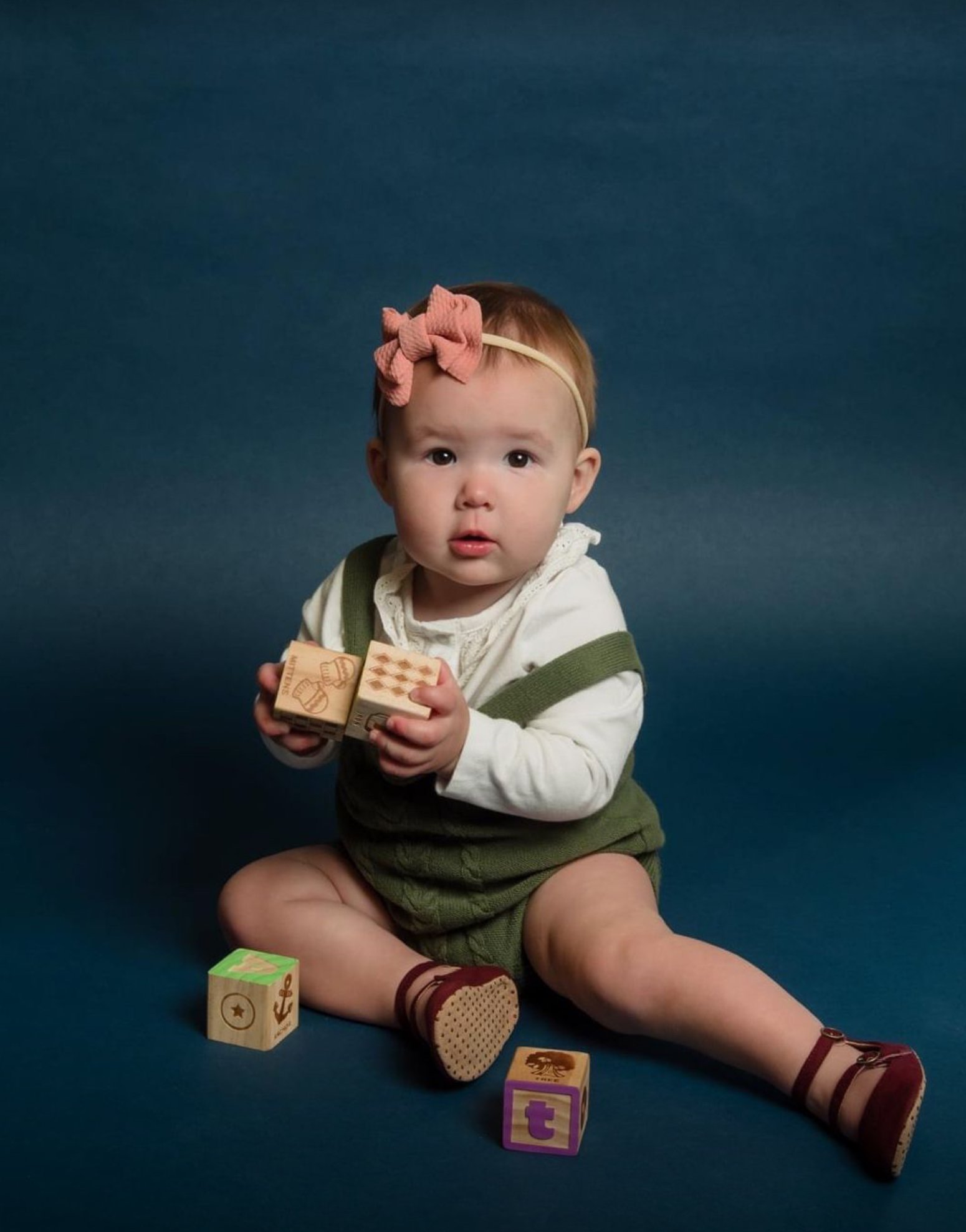 A baby girl sitting on a dark blue background, holding wooden blocks, with additional blocks around her.