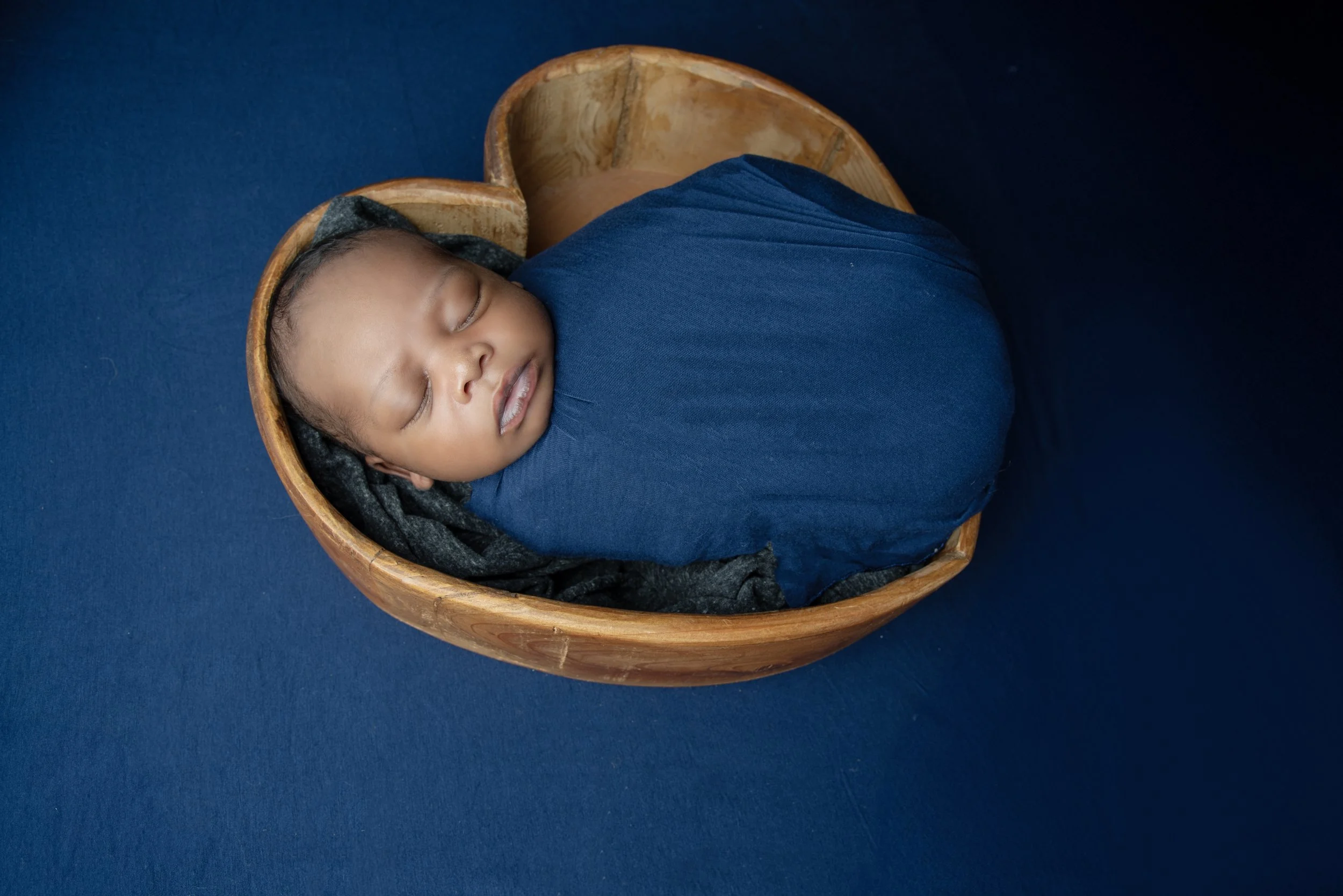 Newborn Baby boy wrapped in blue fabric in heart shaped bowl lying on blue fabric
