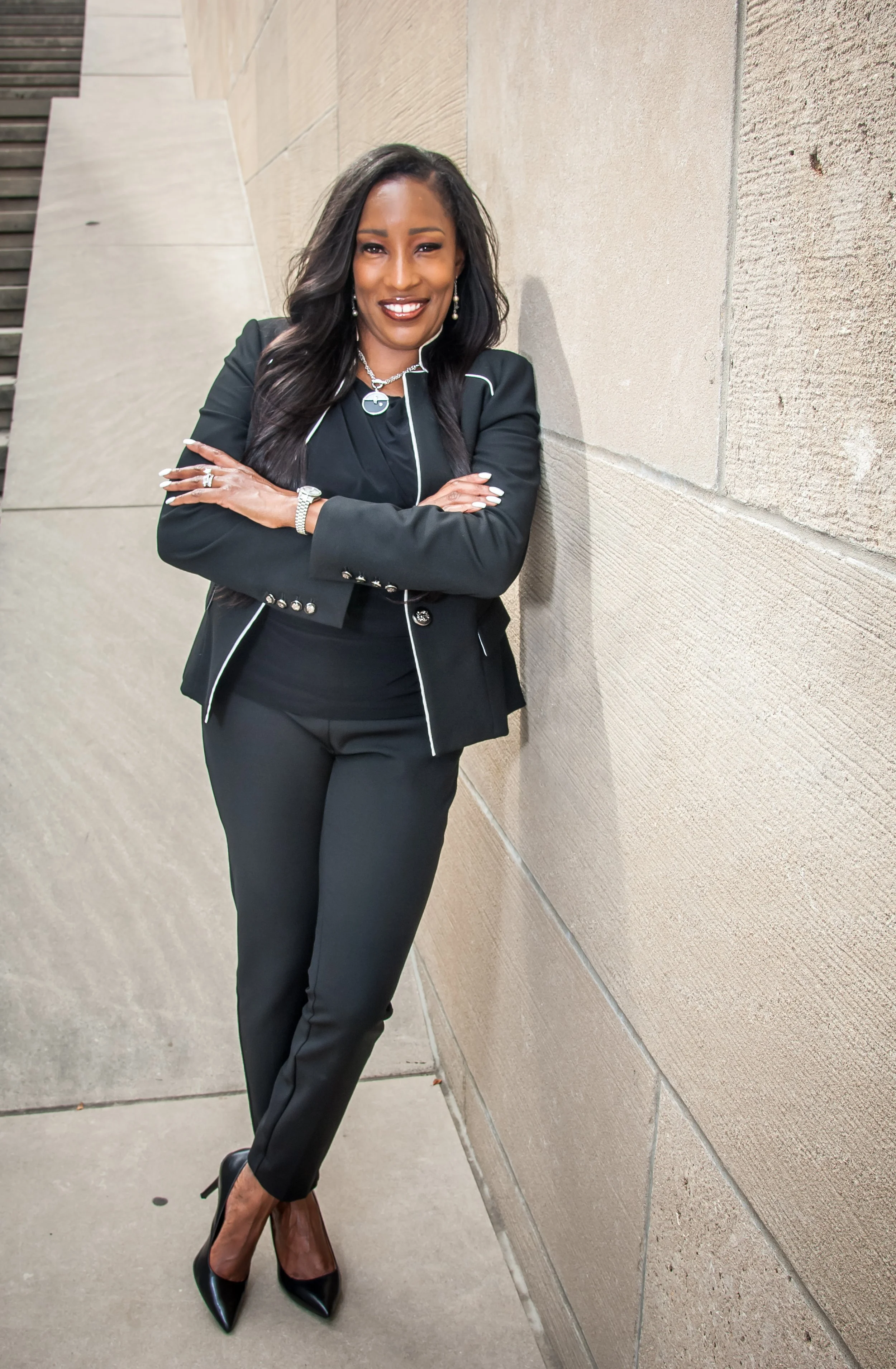 A woman in a black suit with white piping, black heels, and jewelry, standing with her arms crossed, smiling, and leaning against a beige wall outdoors.