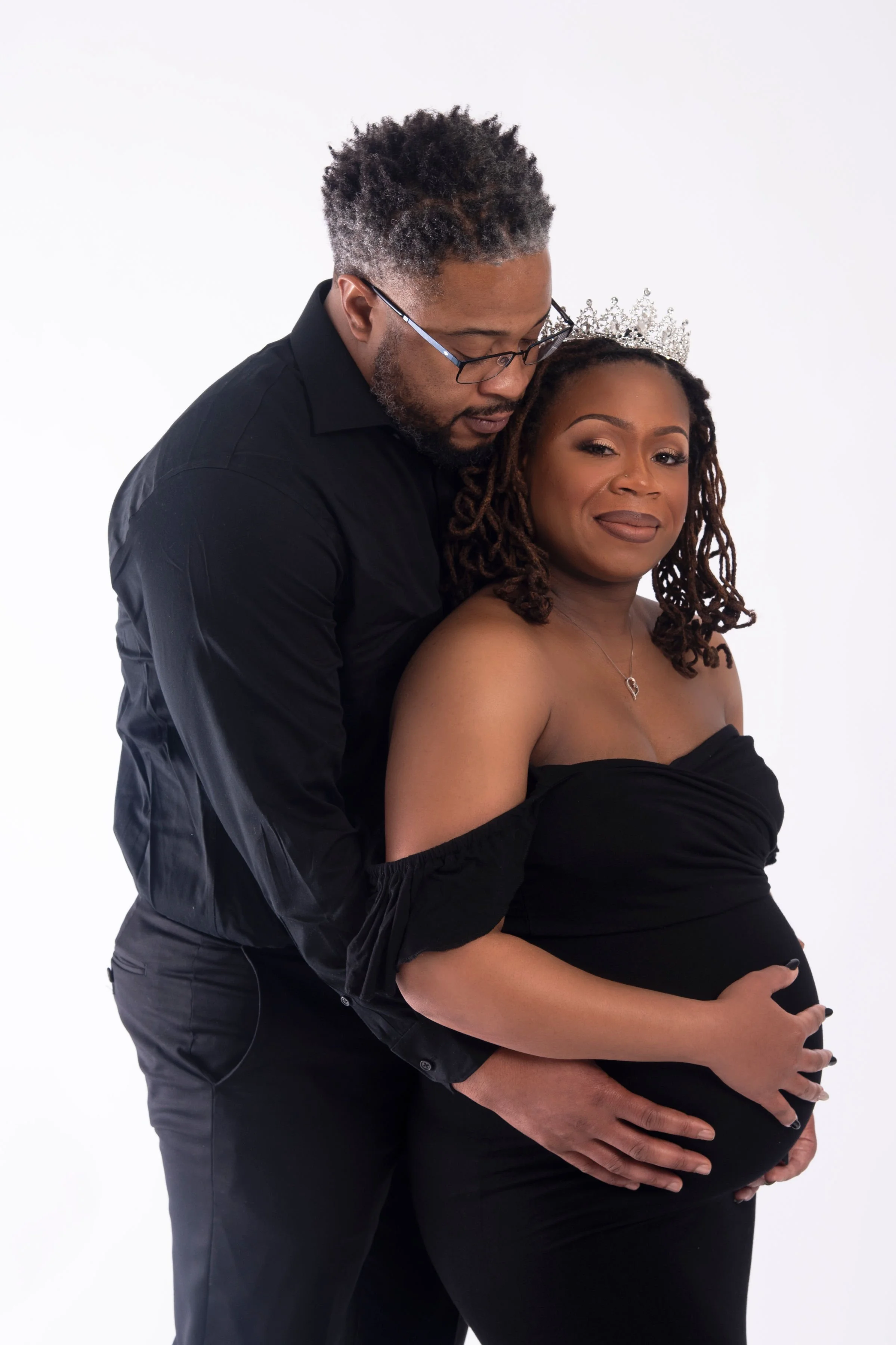 A pregnant woman wearing an off-shoulder black dress and a tiara, with a man in glasses and a black shirt standing behind her, both holding her baby bump against a white background.