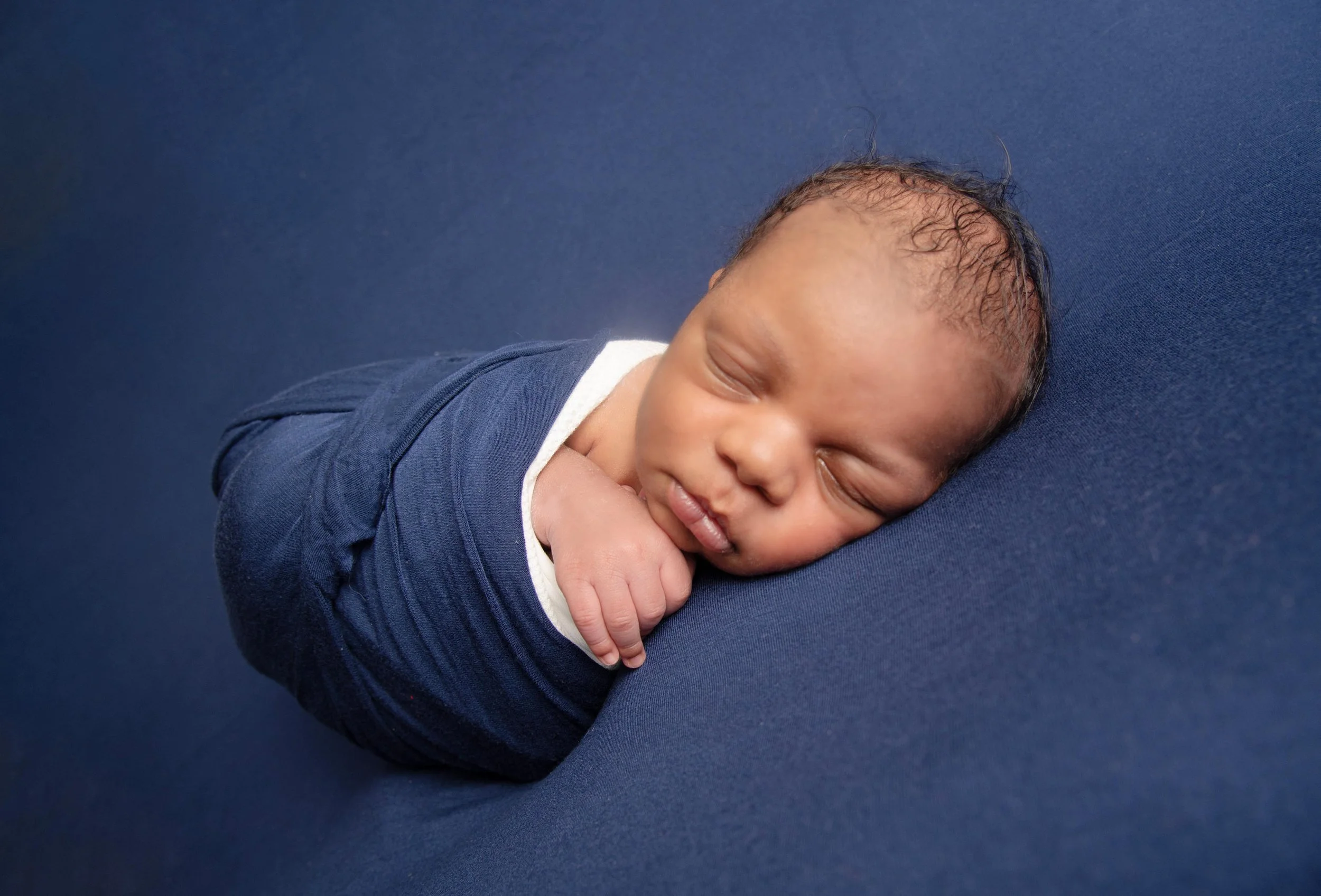 A sleeping newborn baby swaddled in a dark blue blanket and resting on a matching blue surface.