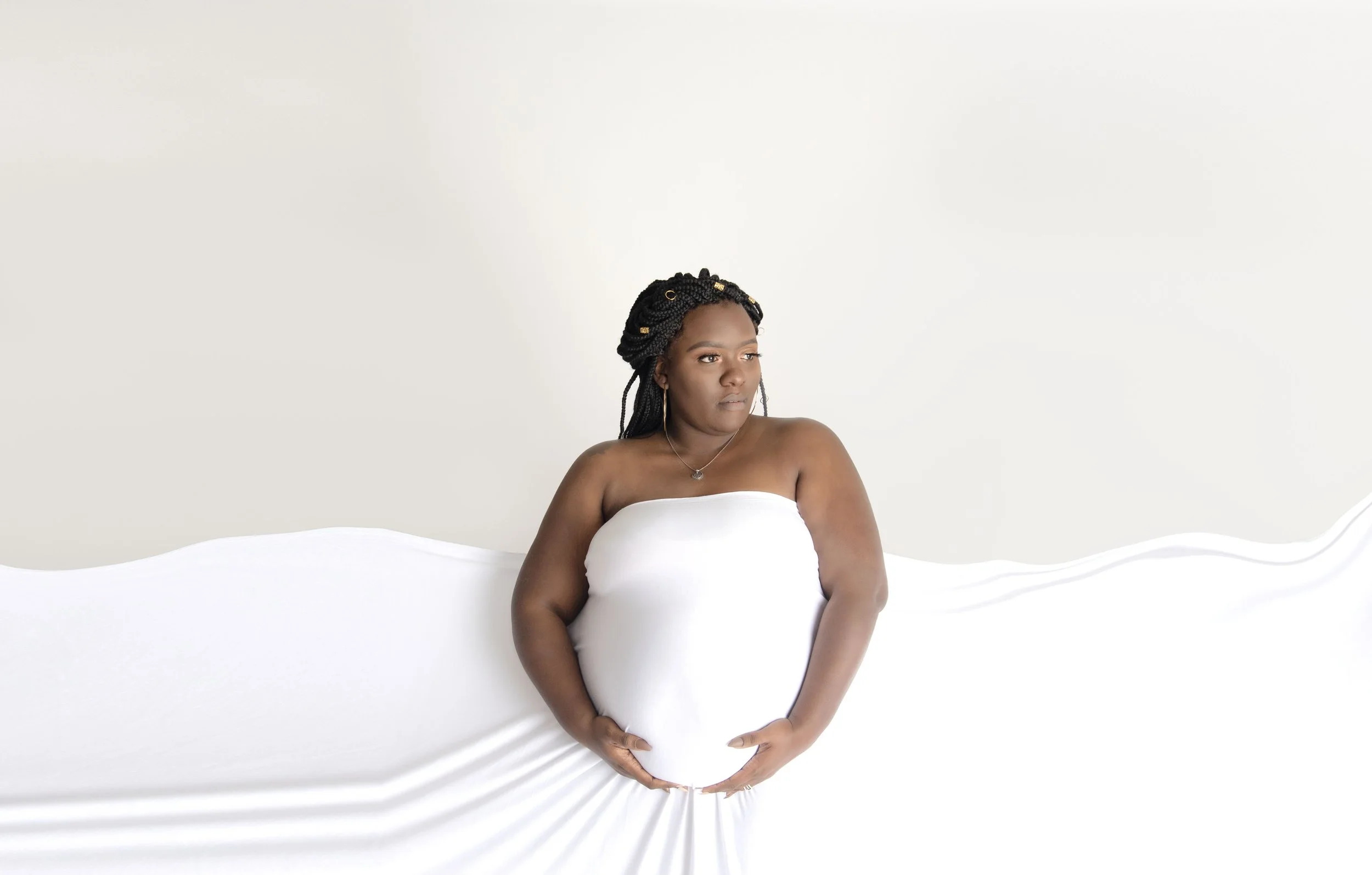 Expecting mama cradling baby bump draped in white fabric against white backdrop