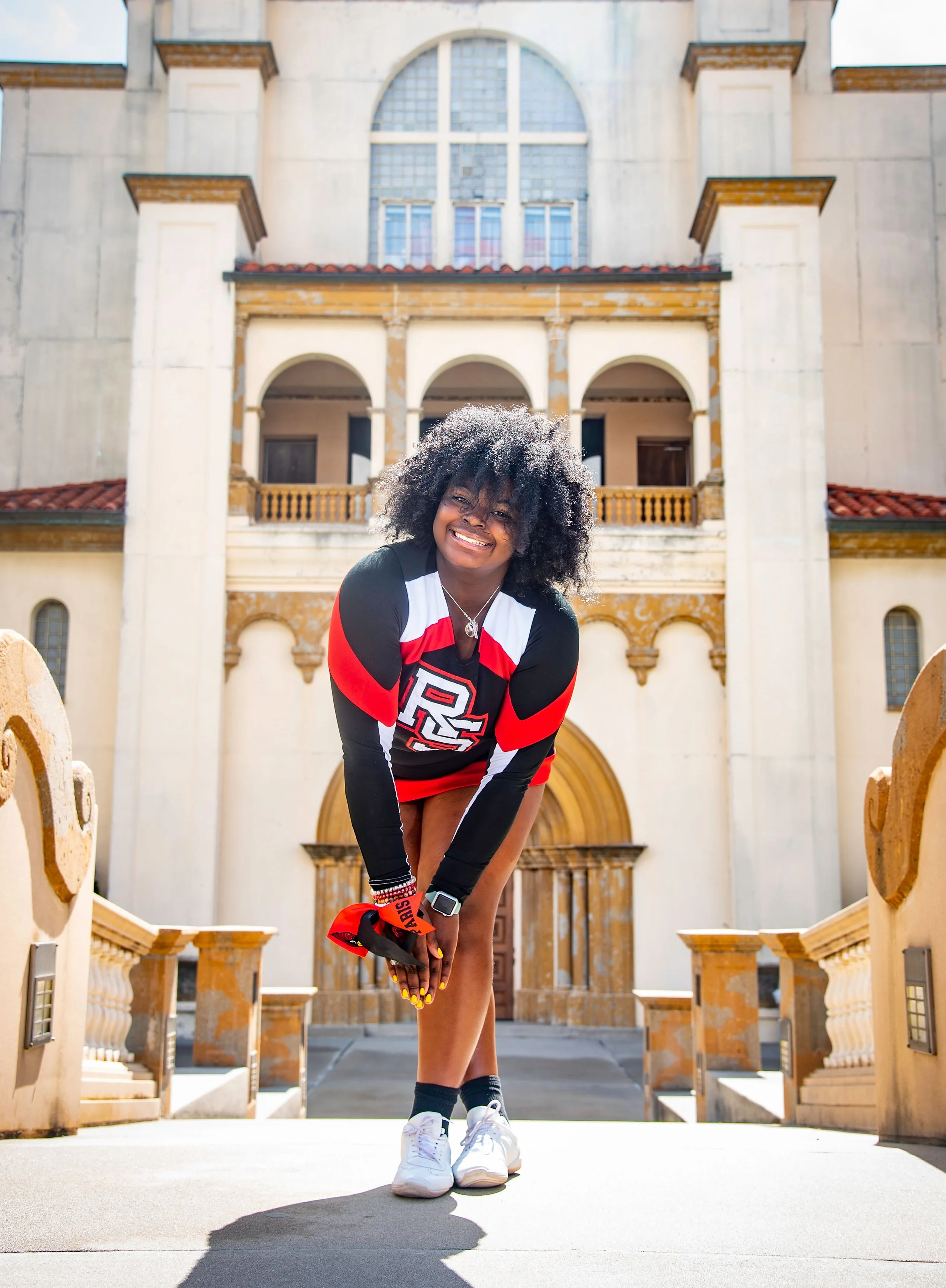 Young woman in cheerleading uniform smiling, standing outside a large historic building with arches and windows, holding her hands together in front of her, with sunlight highlighting her curly hair.