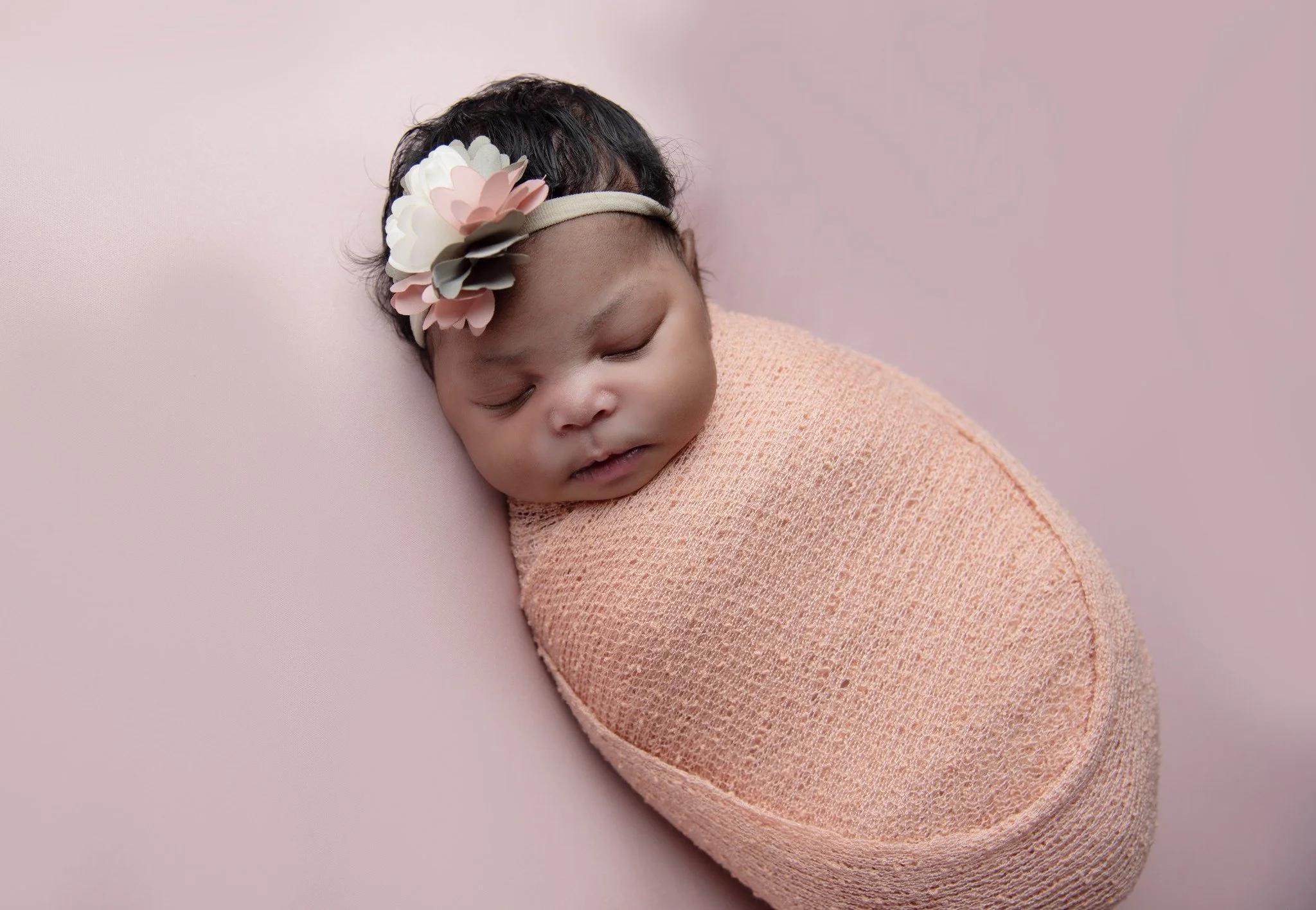 A sleeping baby wrapped in a pink blanket, wearing a flower headband, lying on a pink surface.