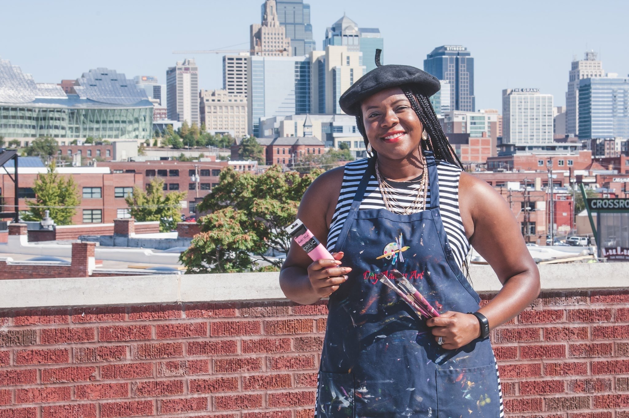 Woman artist smiling outdoors on a rooftop with a city skyline in the background, holding painting supplies and wearing an apron and beret during branding session