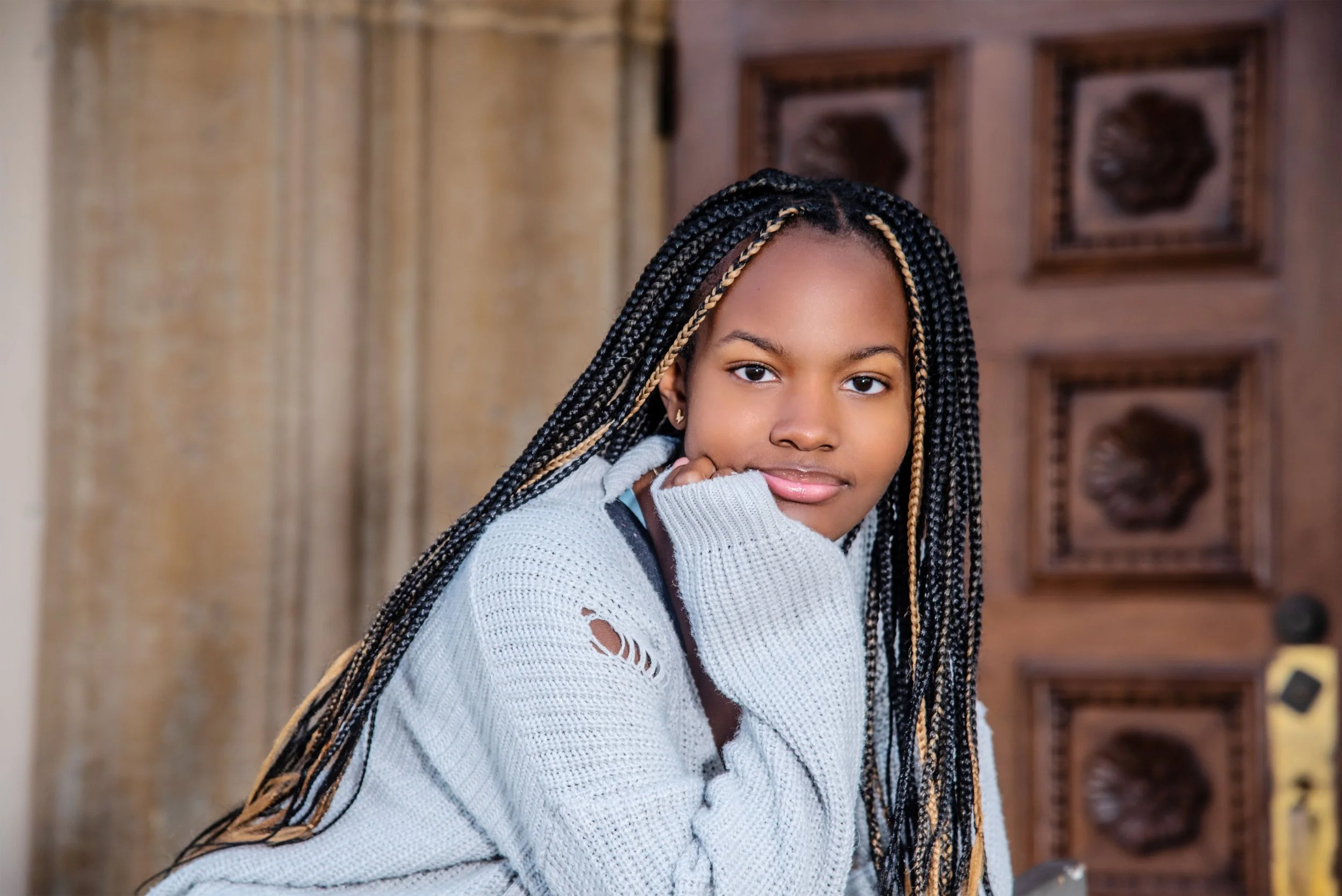 A young woman with braided hair and gold highlights, wearing a gray sweater with small holes, resting her chin on her hand and looking at the camera.