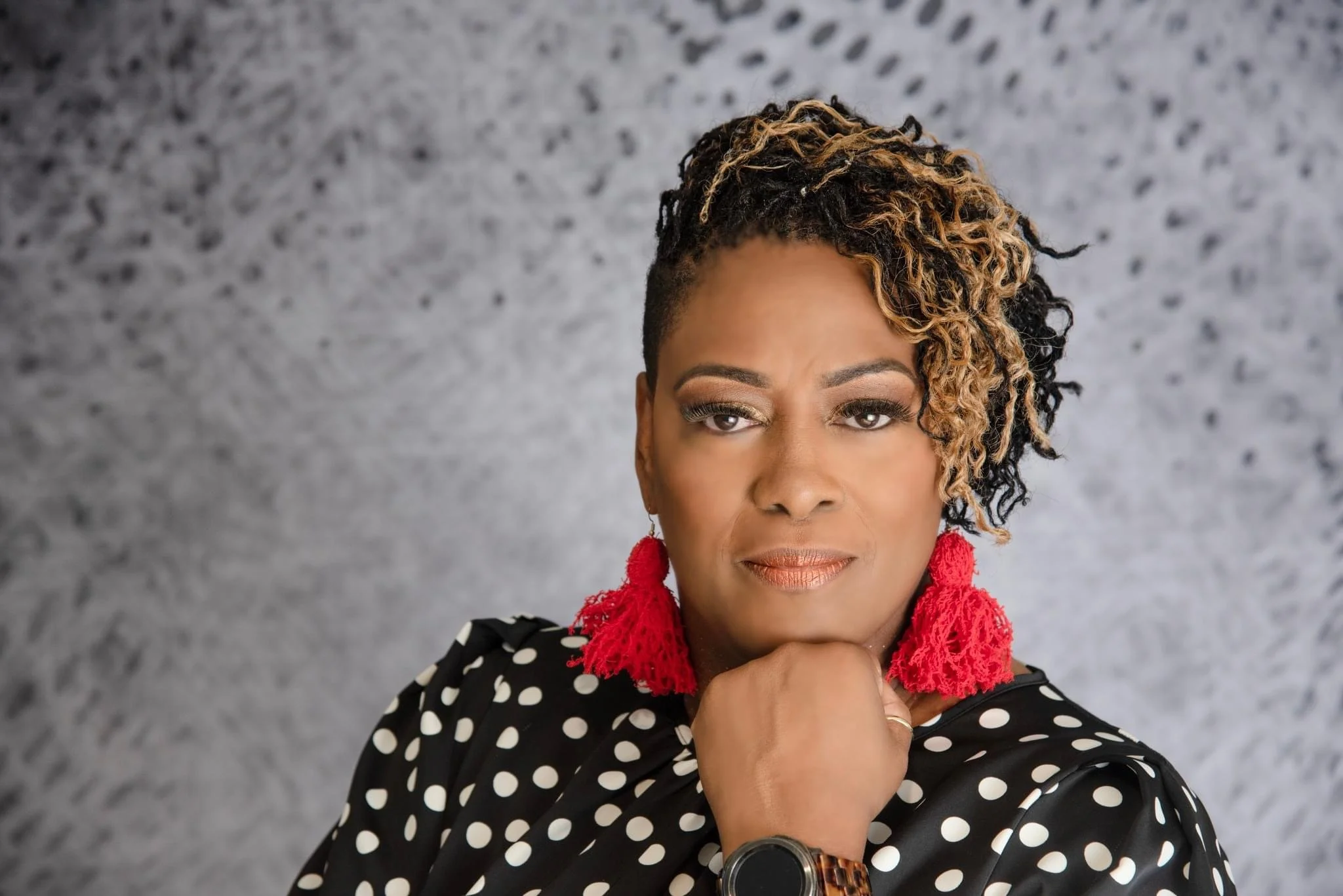 A confident woman with short, styled hair wearing red earrings and a black polka-dot blouse, resting her chin on her hand, against a gray textured background for small business headshot.