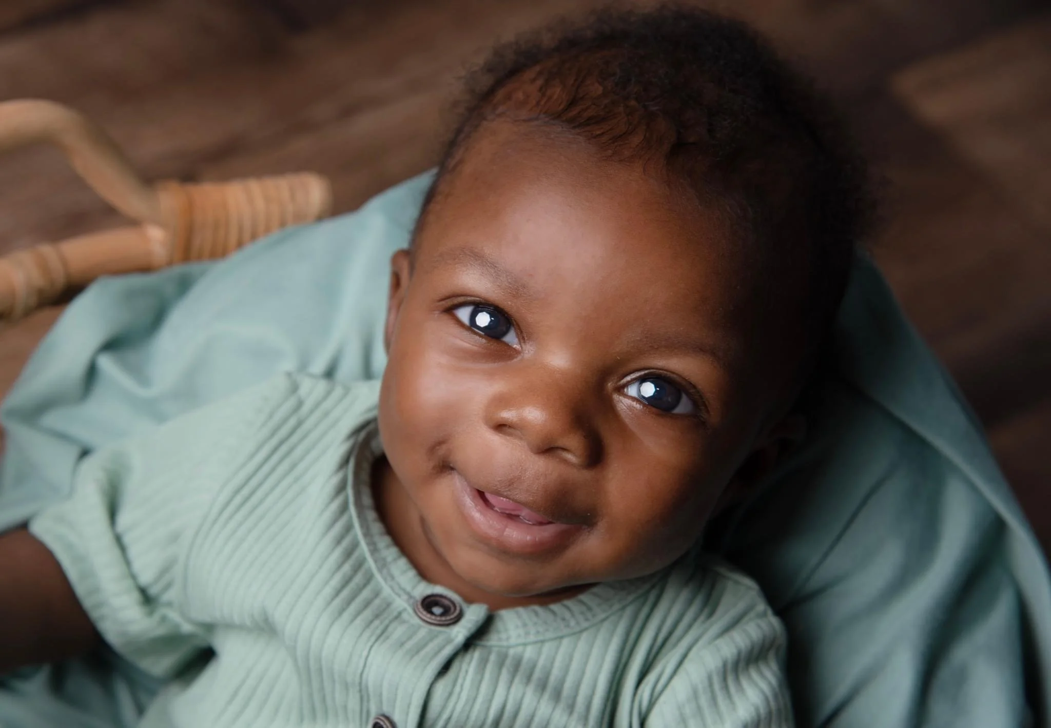 A smiling young African-American child with short curly hair looking up at the camera, wearing a button-up shirt, lying on a blue cushion on a wooden floor.
