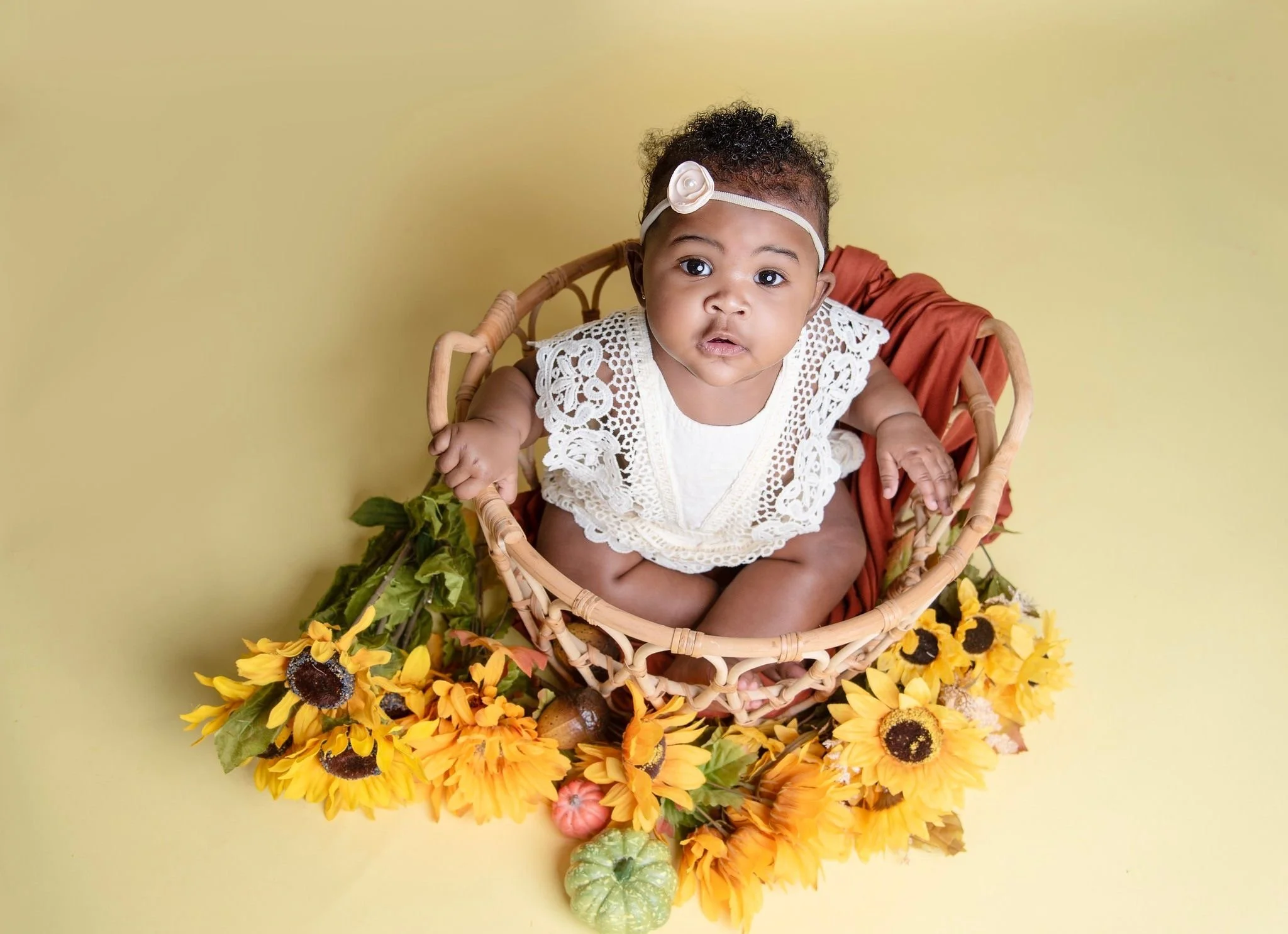 Baby in a woven basket surrounded by sunflowers and other small pumpkins, looking up with curious expression, wearing a white lace top and a headband with a flower.