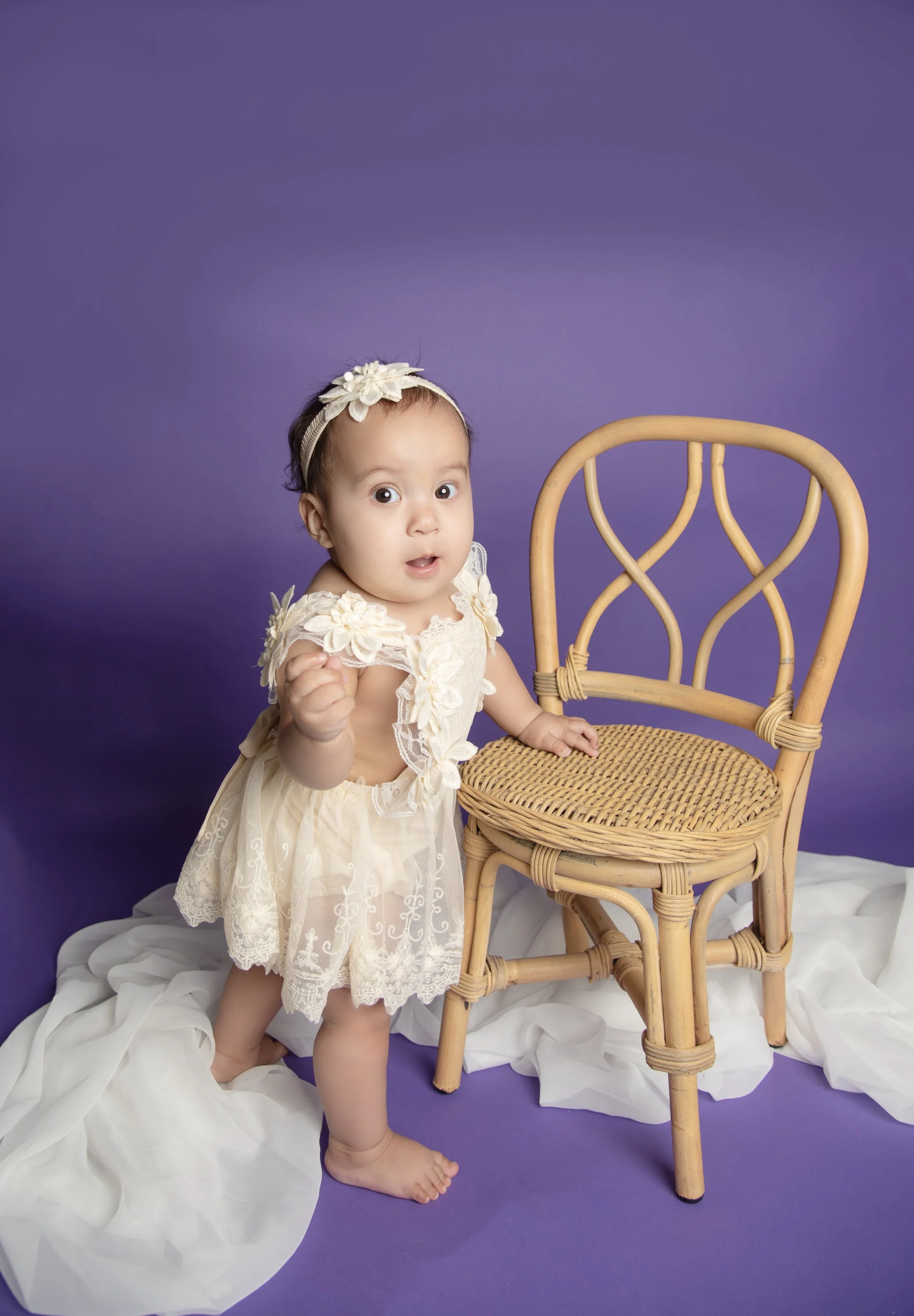 A baby girl with a headband and a cream-colored dress standing barefoot next to a wicker chair on a purple background with white fabric on the floor.