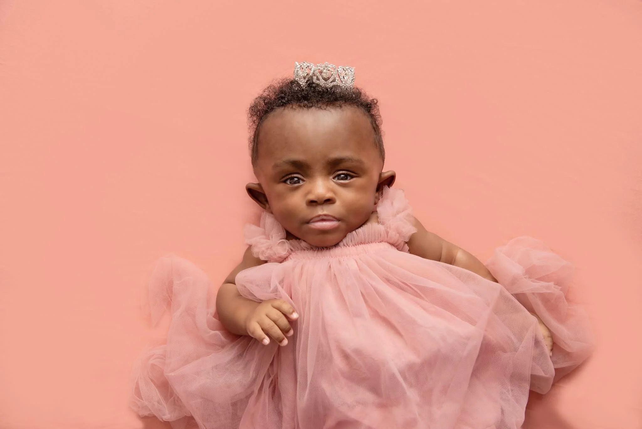 Baby girl with a tiara, wearing a pink tulle dress, lying on a pink background.