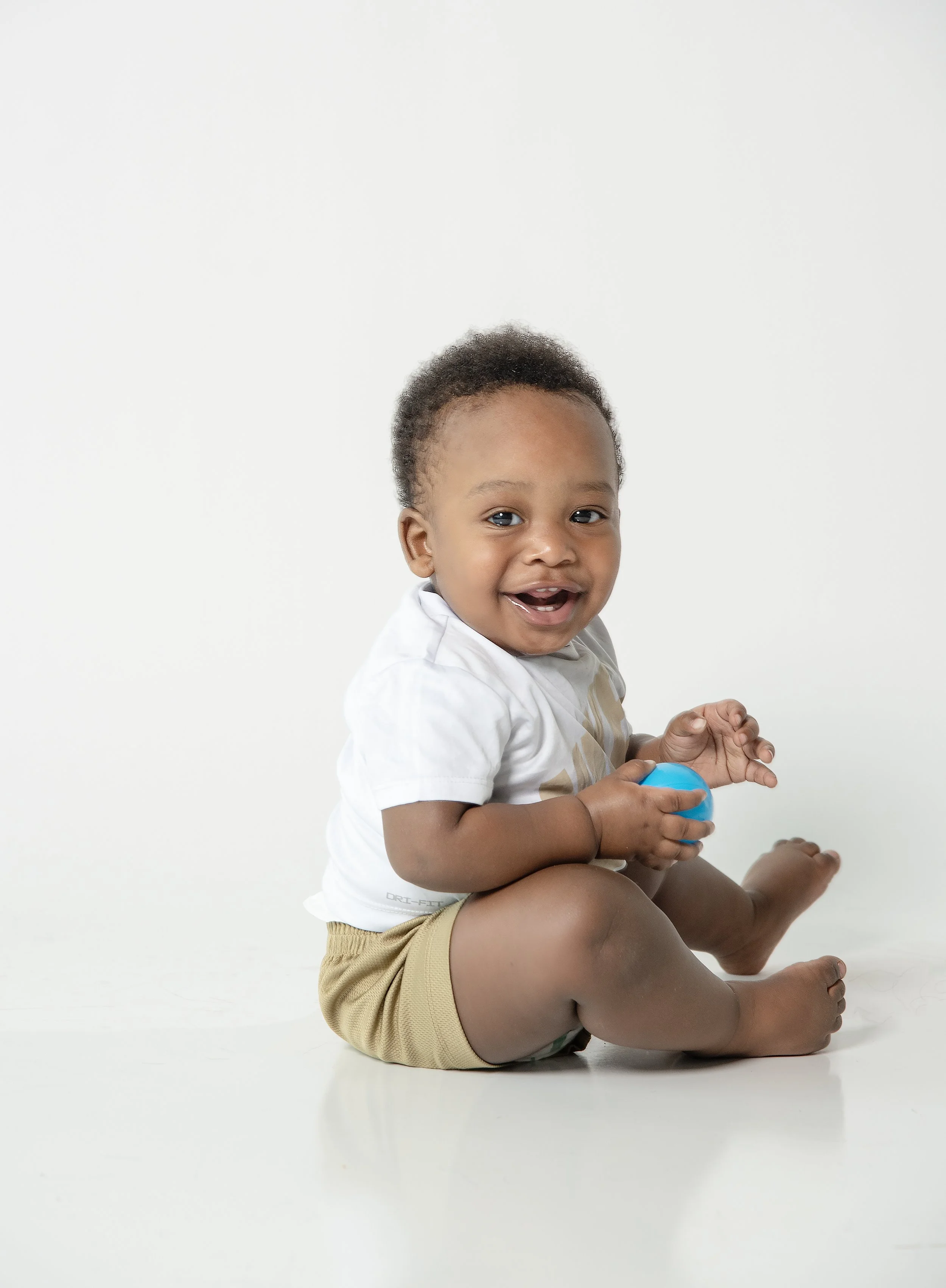 A smiling baby sitting on the floor holding a blue ball, wearing a white shirt and beige shorts, against a plain white background.