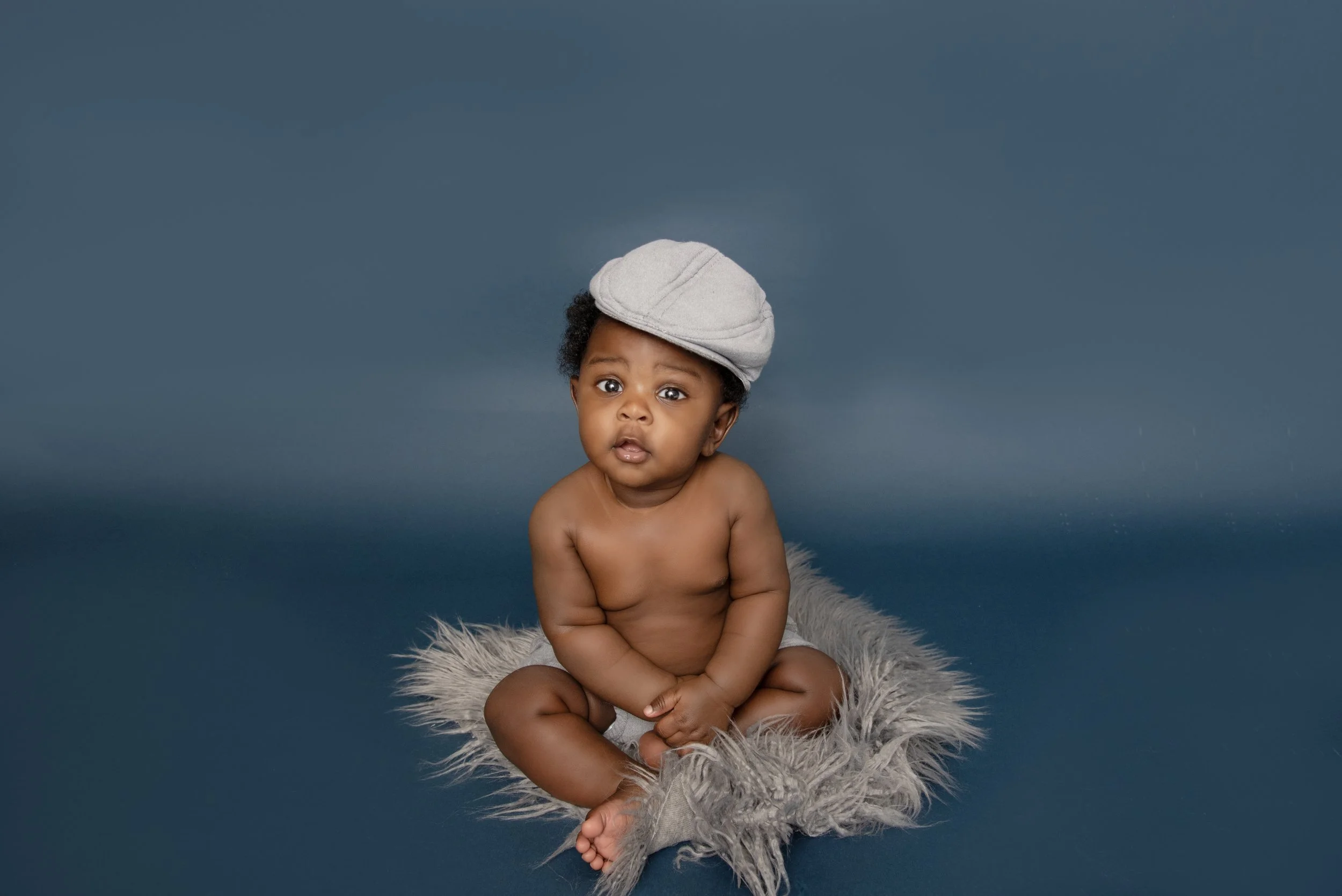 A young baby sitting on a fluffy gray rug, wearing a gray cap, with a surprised expression, against a dark blue background.