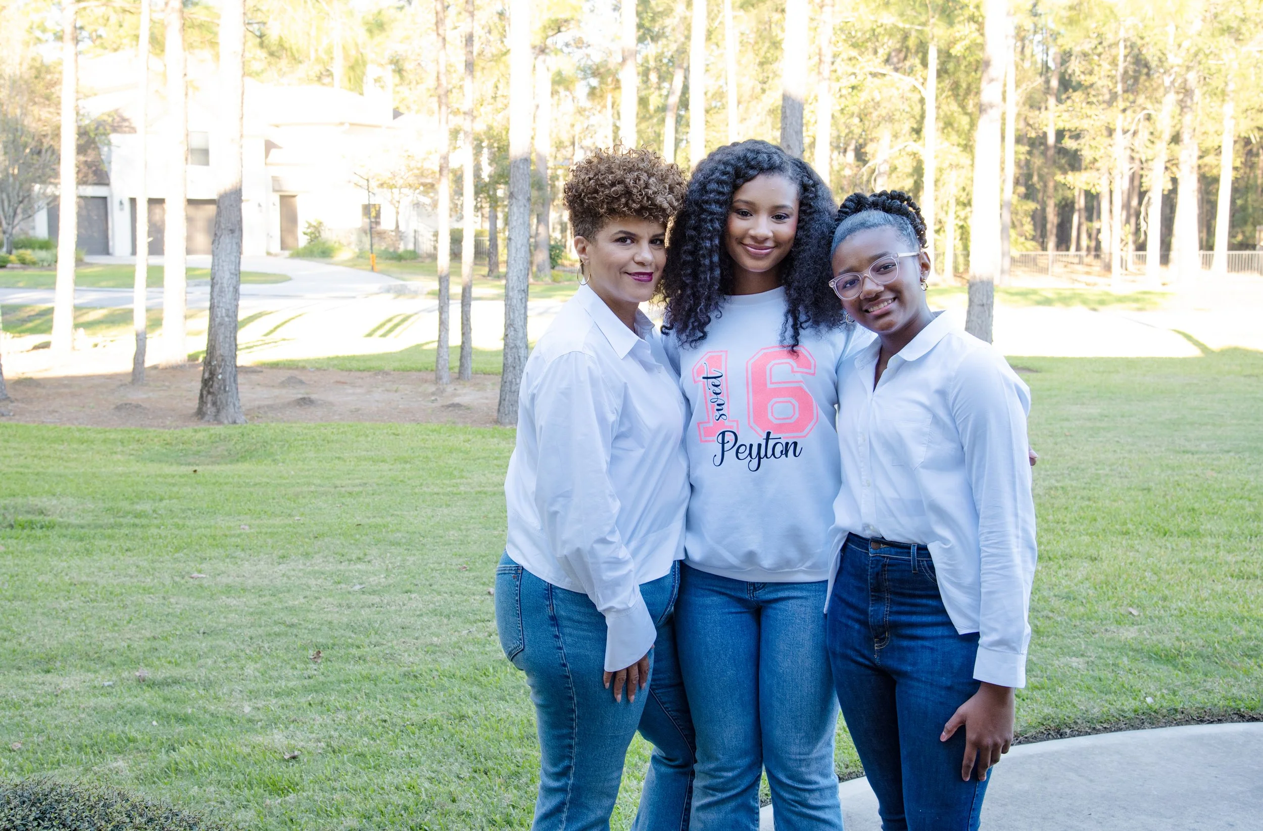 Three women standing together outdoors on a grassy area, smiling at the camera, with trees and houses in the background.