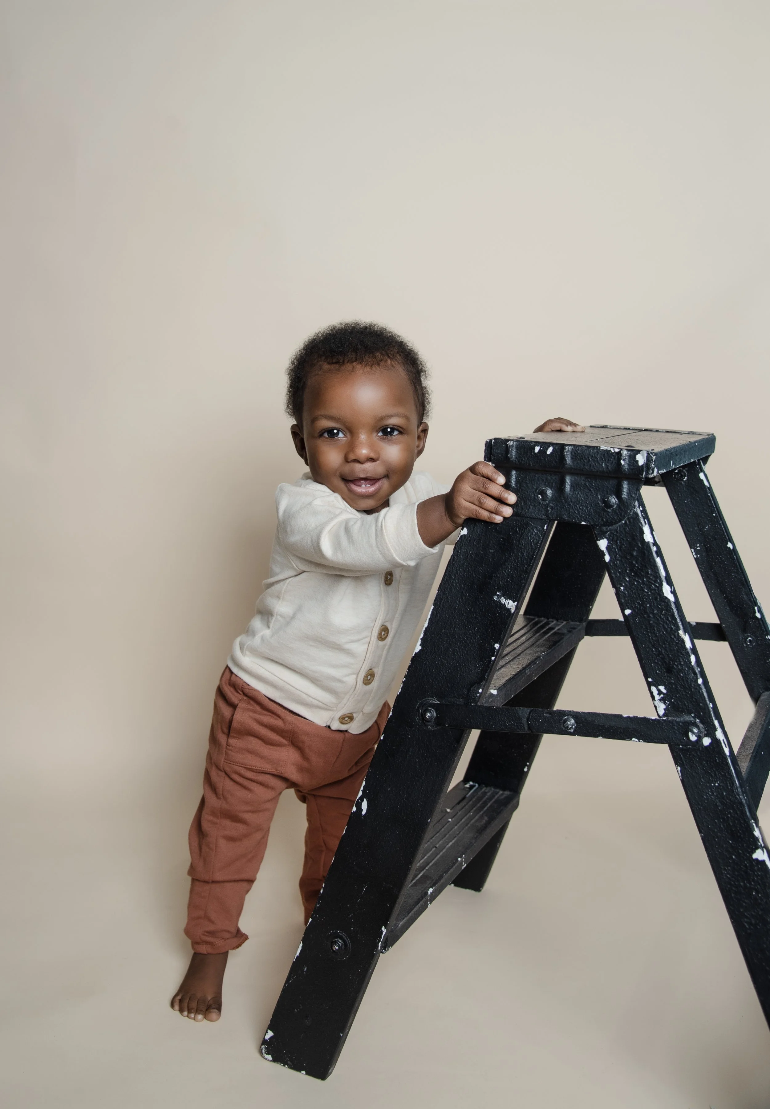 A young child with dark skin, curly hair, wearing a off-white sweater and brown pants, standing beside a black step ladder, holding onto the top of the ladder and smiling.