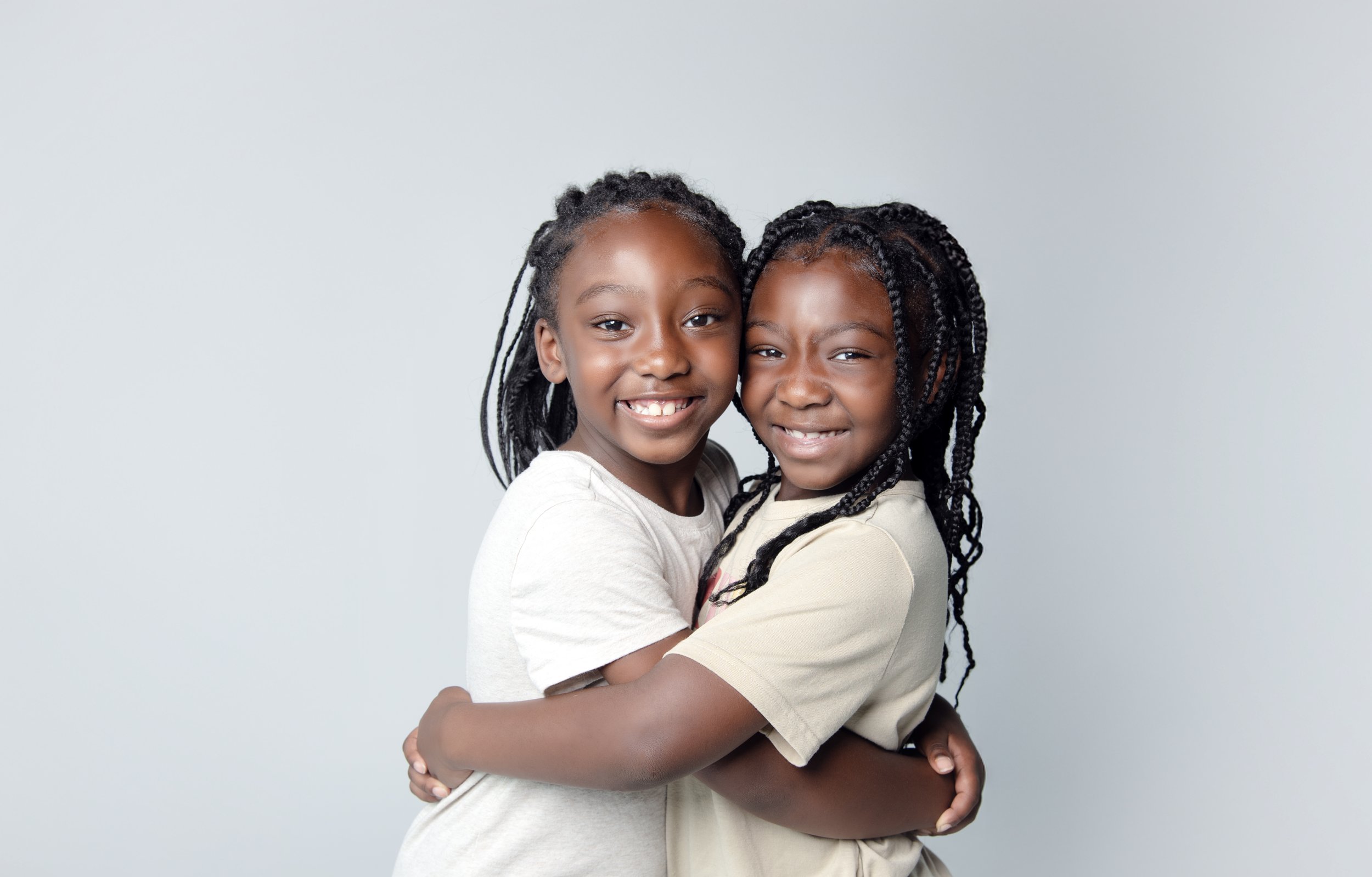 Two young girls with braided hair hugging and smiling at the camera against a plain light gray background.